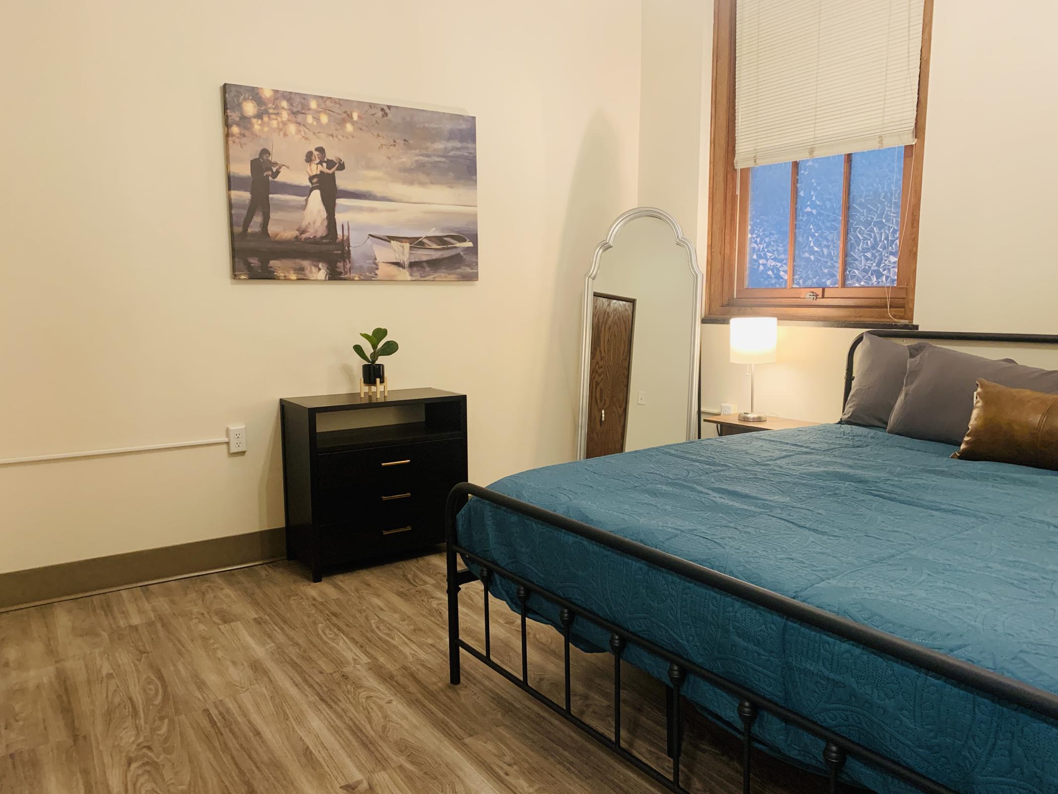 A cozy bedroom featuring a black metal bed with a teal quilt, a stylish nightstand with drawers, and a decorative mirror. A painting of a couple dancing hangs on the wall, and a potted plant adds a touch of greenery. Natural light filters in through a window with closed blinds.