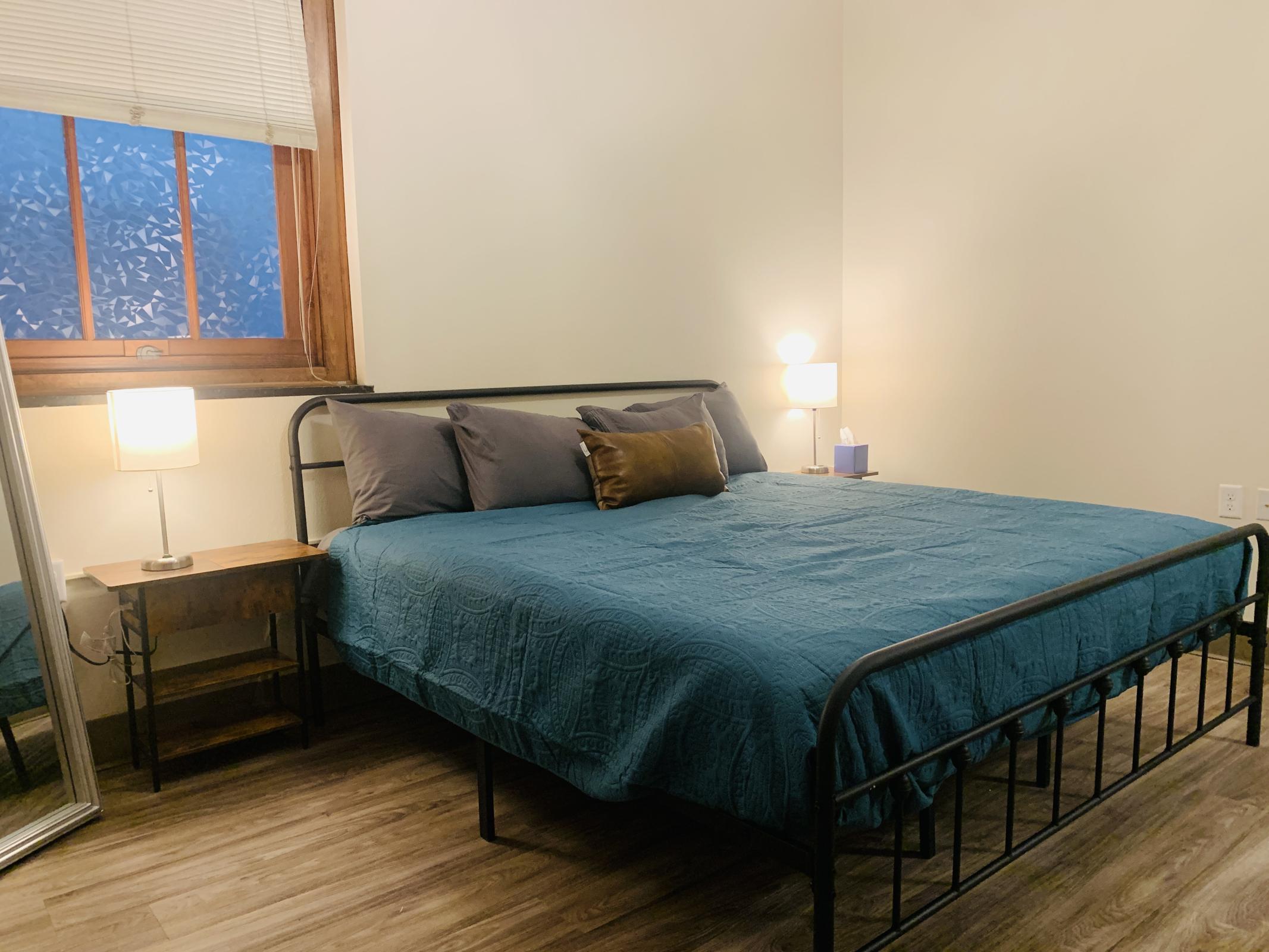 A cozy bedroom featuring a metal frame bed with a teal quilt, two gray pillows, and a brown accent pillow. There's a nightstand with a lamp on one side and a full-length mirror on the other. Natural light filters in through a window, creating a warm atmosphere. The flooring is wooden.