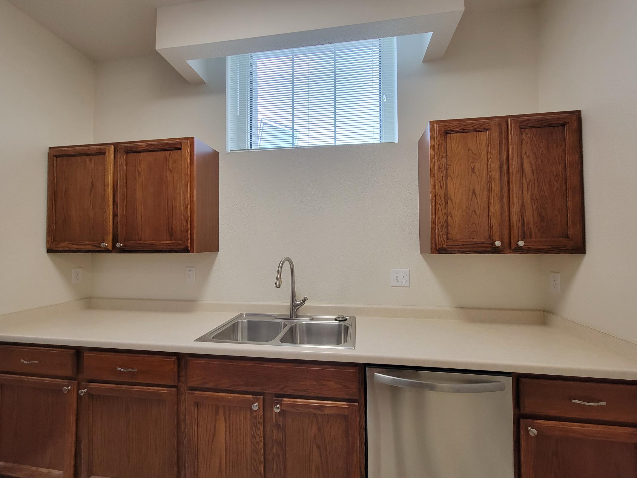A modern kitchen featuring light-colored walls and cabinetry. The countertop is beige, with a double stainless steel sink below a window. Above the sink are two wooden cabinets on either side, and a stainless steel dishwasher is seen on the right. The space is bright and uncluttered.
