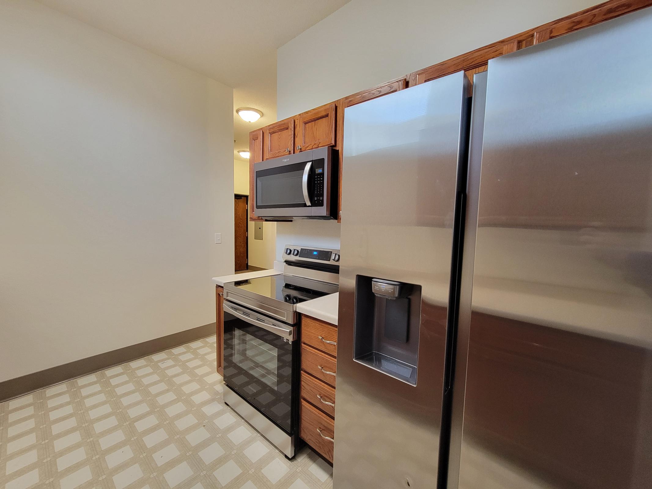 Modern kitchen featuring stainless steel appliances including a refrigerator, oven, and microwave. The cabinetry is wooden with a clean countertop, and the floor has a light checkered pattern. Natural light floods the space, creating an inviting atmosphere.