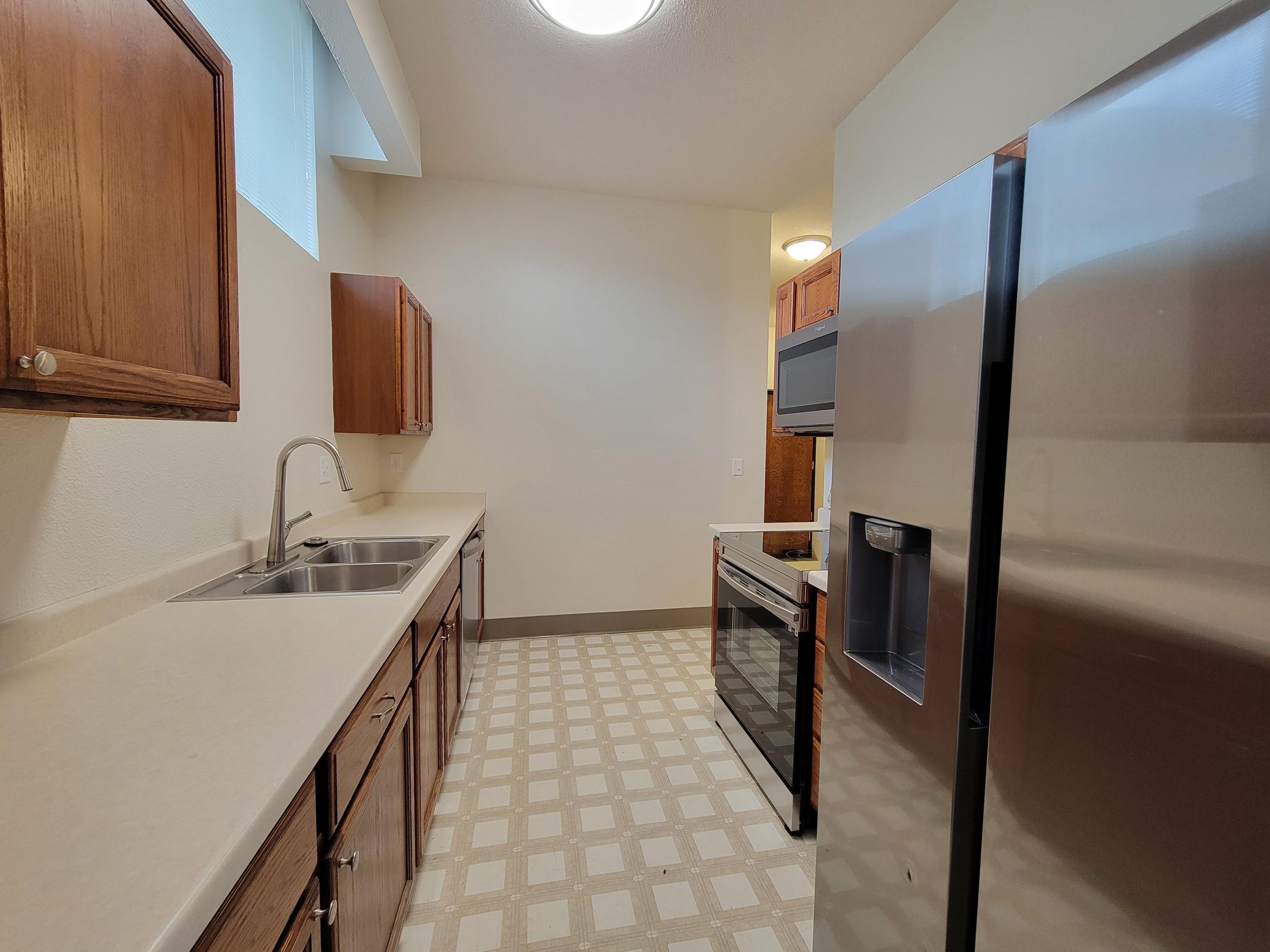 A modern kitchen featuring light-colored walls and cabinetry, stainless steel appliances, and a large window providing natural light. The layout includes a long countertop, double sink, refrigerator, and an oven with a stovetop. The floor has a checkered pattern with light and dark squares.