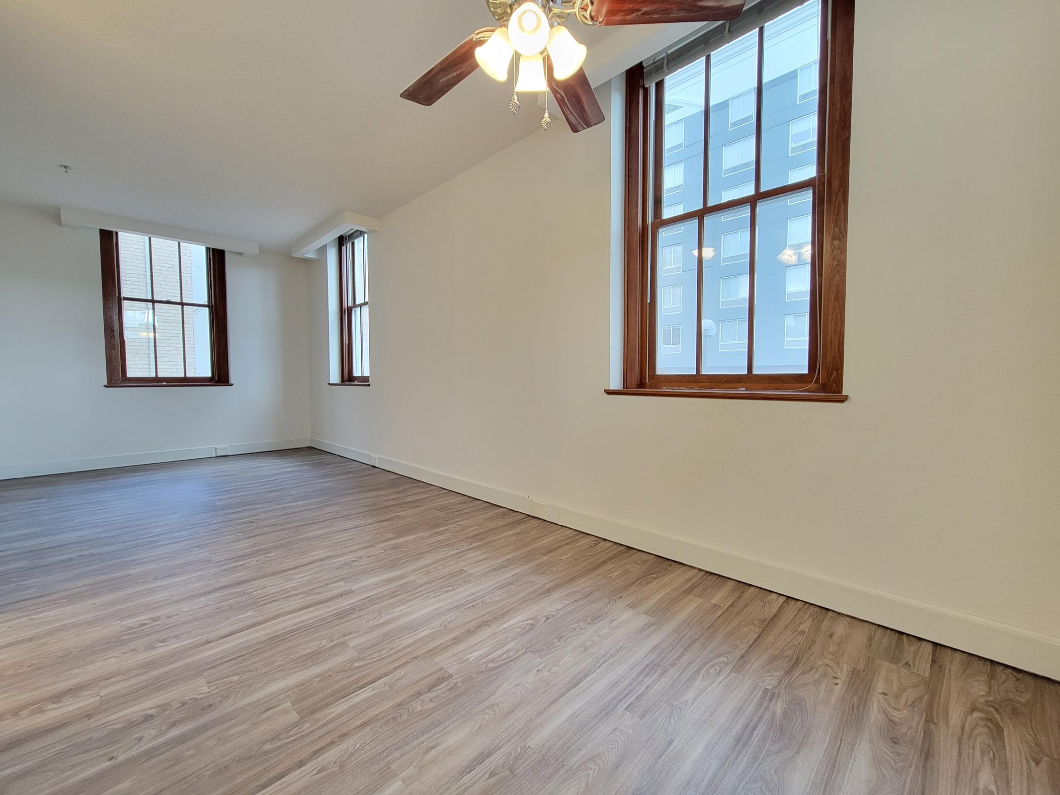 Spacious empty room with light-colored walls and large windows allowing natural light. Features wooden flooring and a ceiling fan with three blades. The room is unfurnished, creating an open and airy atmosphere, ideal for customization.