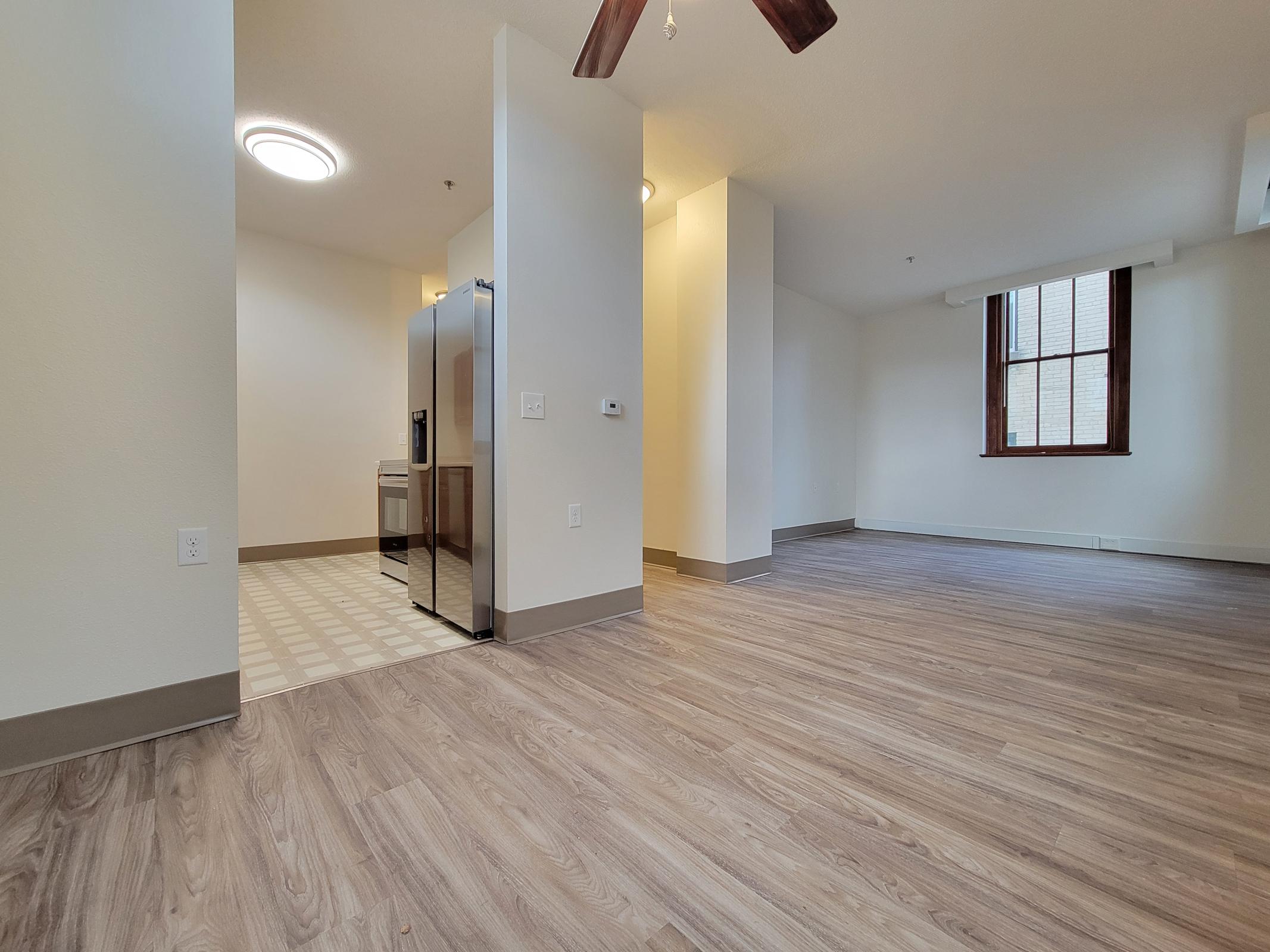 Spacious interior of an apartment featuring light-colored walls, a wooden floor, and a kitchen area visible in the background. A stainless steel refrigerator is present, along with a ceiling fan in the living space. Large windows provide natural light, creating an inviting atmosphere.