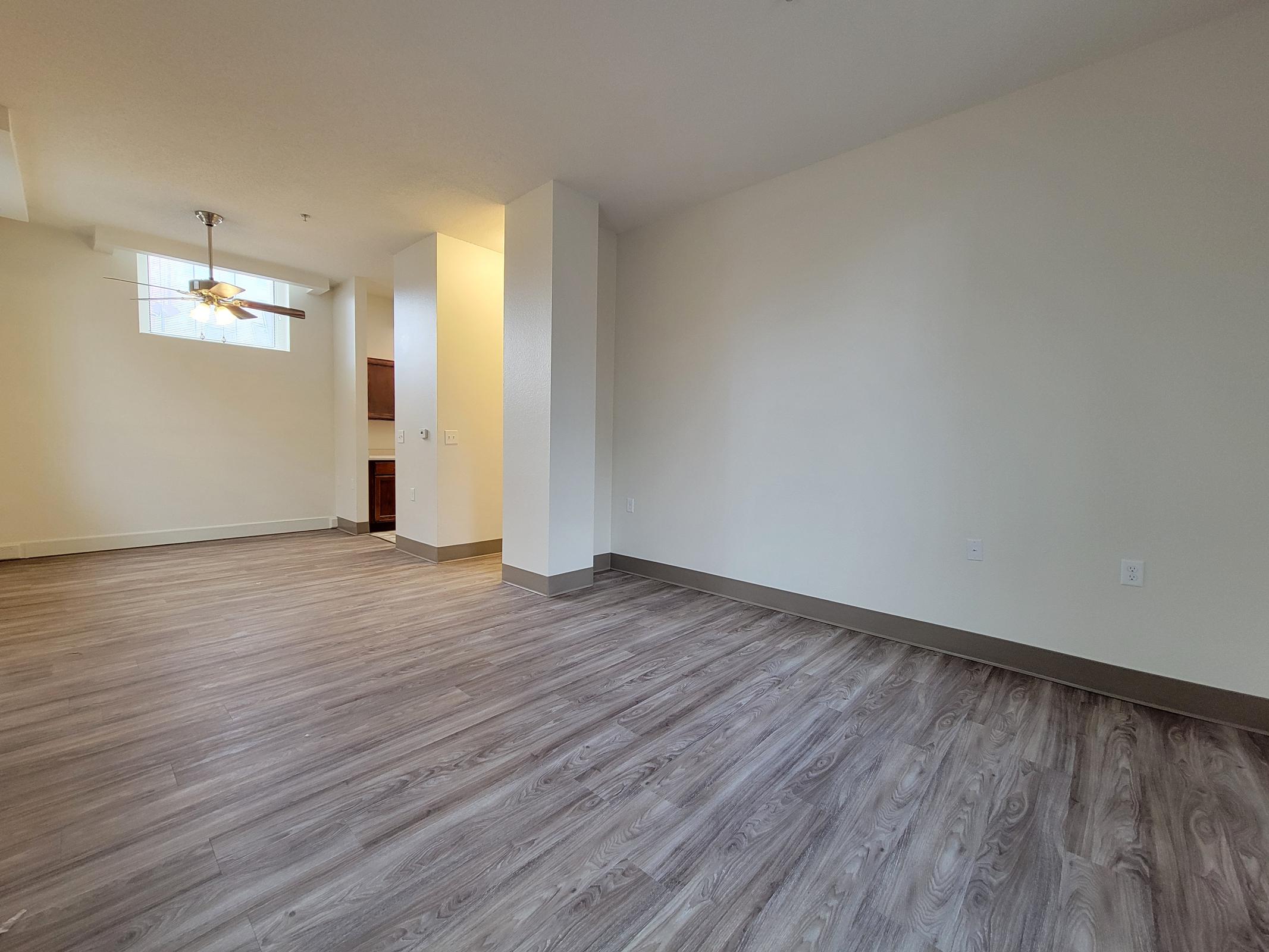 A spacious, empty room featuring light-colored walls and a wood-like floor. There is a ceiling fan and a small window allowing natural light. One corner shows a kitchen area with cabinetry. The room is designed for versatile use, making it suitable for living or dining space.