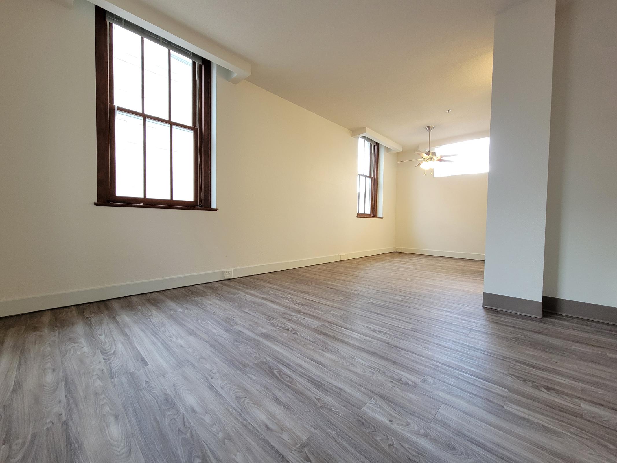 A spacious, empty room with light-colored walls and wooden floors. Two large windows let in natural light, and a ceiling fan is visible in the corner. The room has a modern design with minimal furnishings, creating a bright and airy atmosphere.