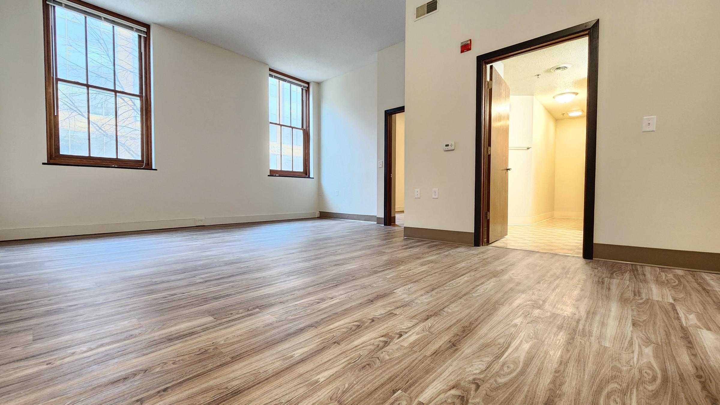 Spacious interior of a room with hardwood-style flooring, large windows allowing natural light, and a doorway leading to a bathroom area. The walls are painted in a neutral color, creating a bright and inviting atmosphere.