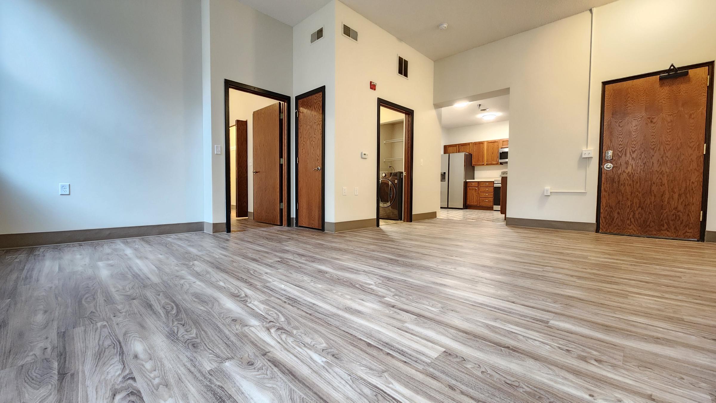 A spacious room with light wood-like flooring, featuring doorways leading to adjacent rooms and a kitchen area visible in the background. The walls are painted in neutral colors, and there is ample natural light coming from a window. The overall atmosphere is modern and inviting.
