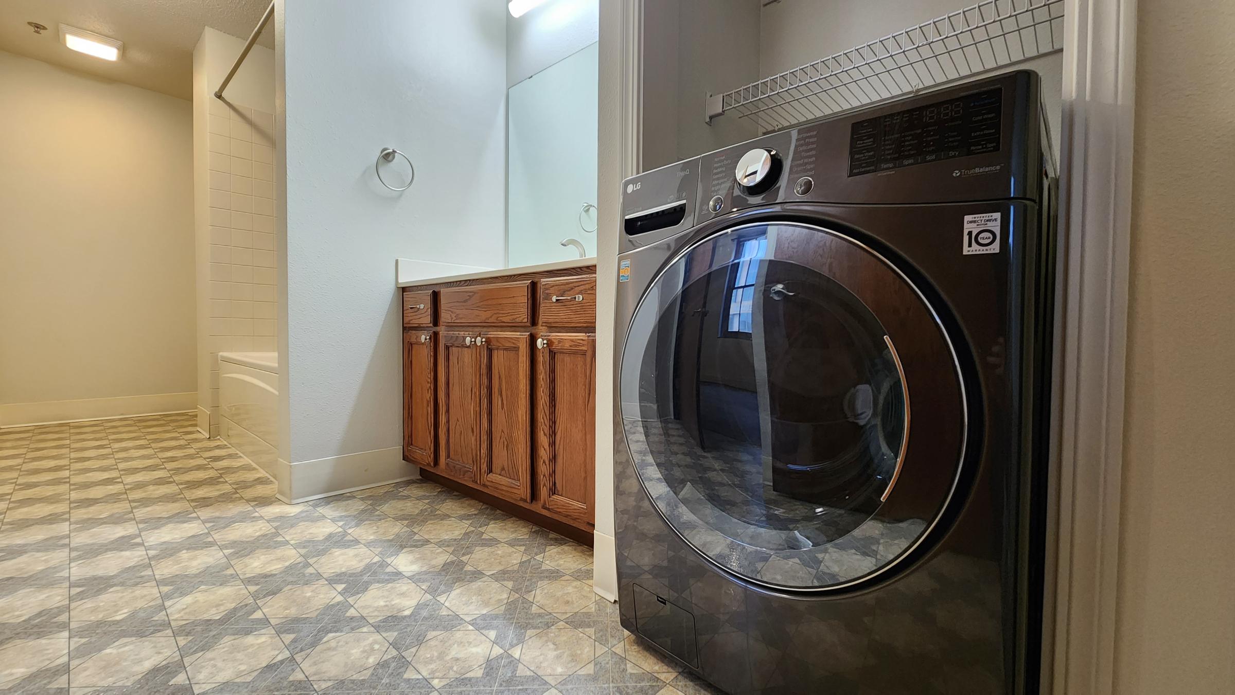 A laundry room featuring a modern front-loading washing machine on the left, with a wooden cabinetry and a countertop visible. The space includes a bathroom area in the background with a sink and mirror, and a shower area partially visible. The floor has a diamond pattern in muted colors.