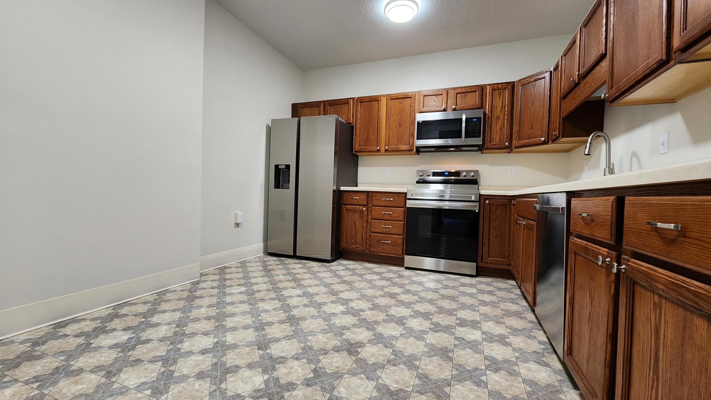 Modern kitchen featuring wooden cabinets, stainless steel appliances including a refrigerator and oven, and a tiled floor with a geometric pattern. The layout is spacious with ample counter space and a microwave above the stove. The walls are painted a light color, enhancing the room's brightness.