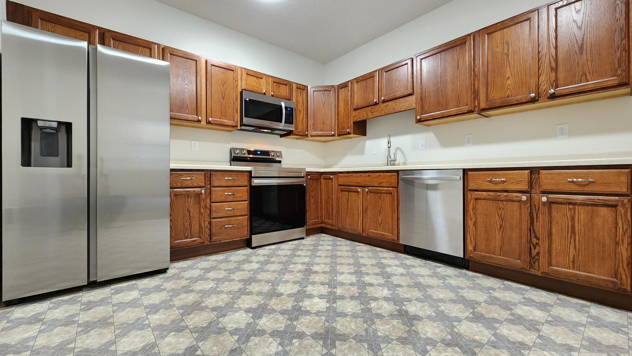Modern kitchen featuring wooden cabinets, stainless steel appliances including a refrigerator, microwave, and oven. The countertop is light-colored, and there's a stainless steel dishwasher. The floor has a tiled pattern in shades of gray and beige, creating a warm and inviting atmosphere.