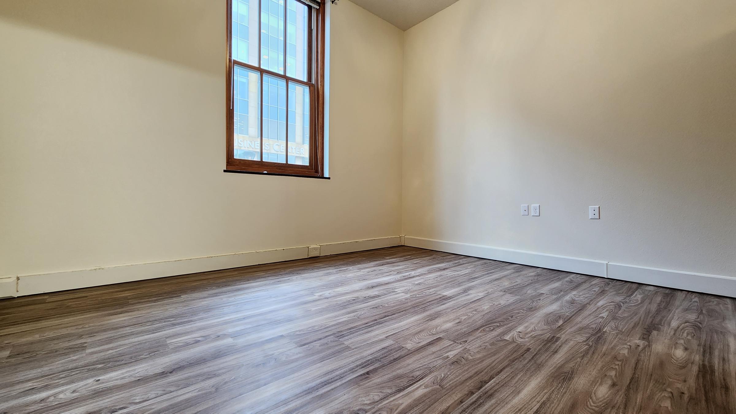 Empty room with light-colored walls, a large window allowing natural light, and modern wood-like flooring. The room features minimal decor, creating a spacious and clean environment.
