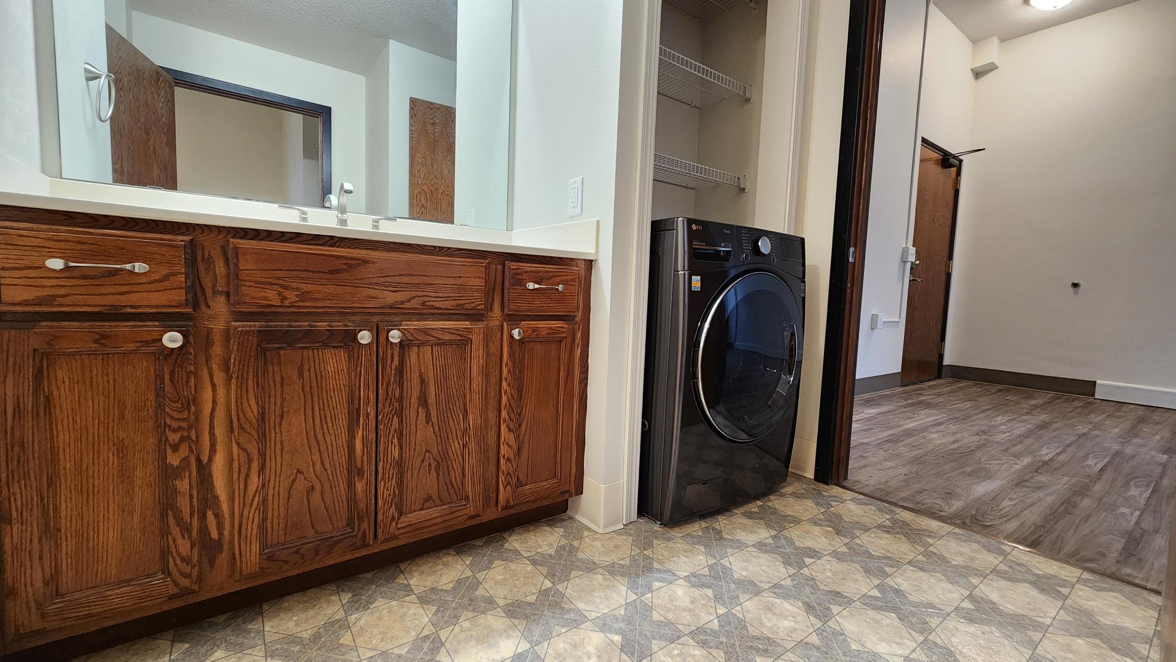 A laundry area featuring a modern black washing machine next to wooden cabinetry with white countertops. The floor has a patterned tile design, and there's an open door leading into a hallway with wooden flooring and a mirror in the background.