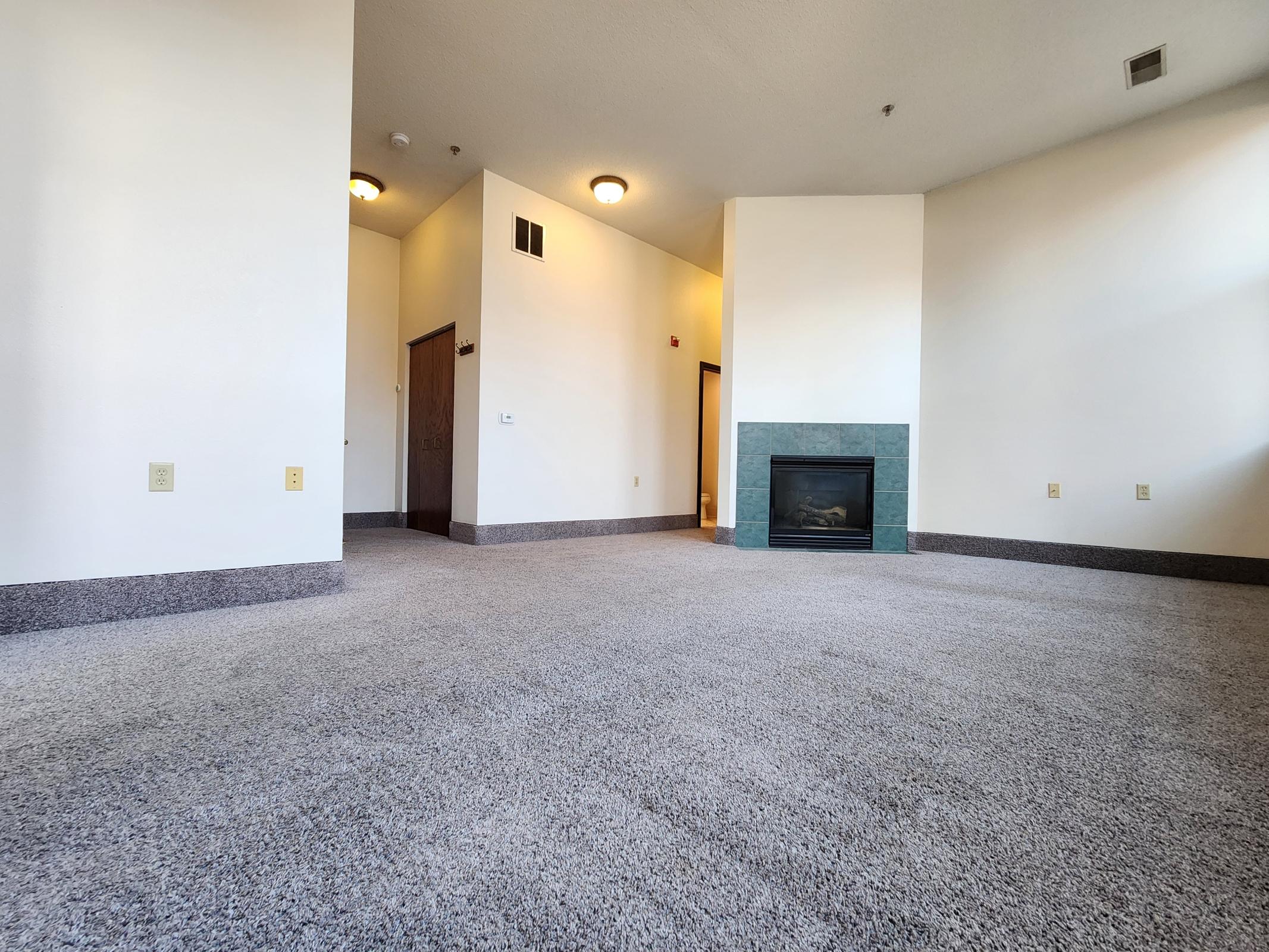 A spacious, empty living room with light-colored walls and a large carpeted floor. To the left, there are doorways leading to other rooms. A green-tiled fireplace is located against the far wall, providing a focal point in the otherwise bare space. Natural light filters in from the windows.