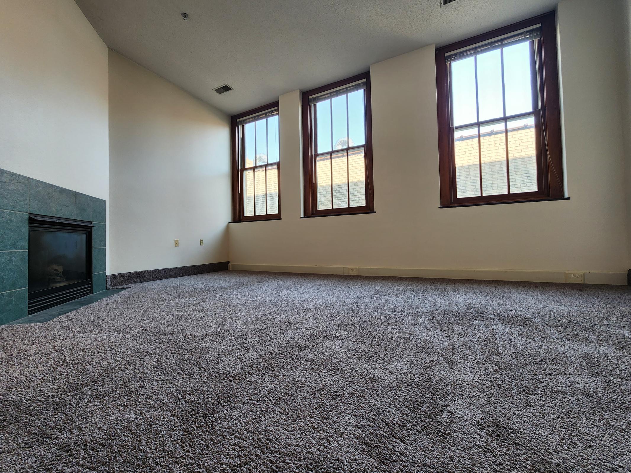 A spacious, unfurnished room featuring three large windows with natural light streaming in. The floor is covered in plush carpet, and there is a simple fireplace along one wall. The overall ambiance is clean and inviting, perfect for personalization and decoration.