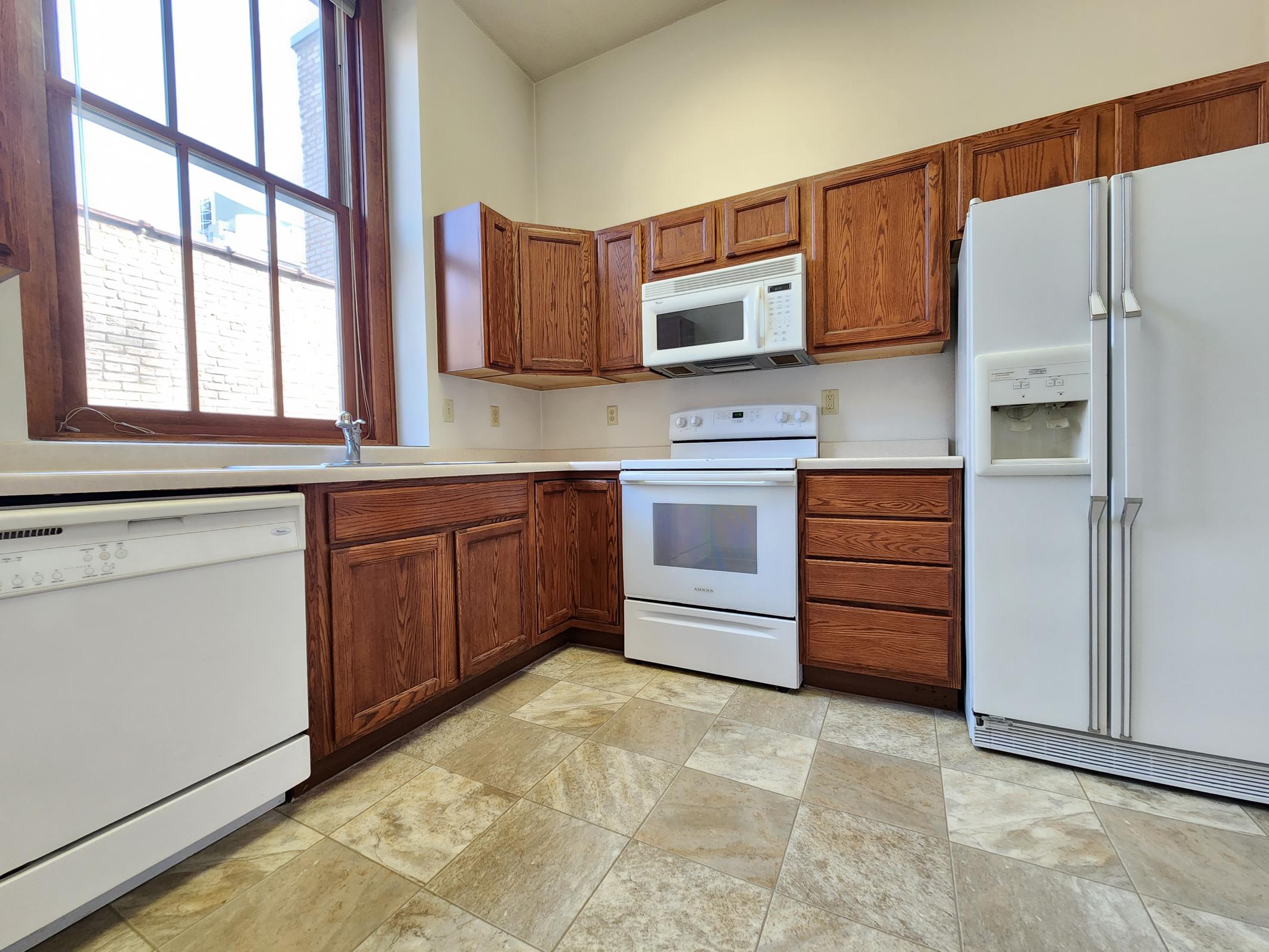 A bright kitchen featuring wooden cabinets, a white refrigerator, a stove, a microwave, and a dishwasher. The room has large windows allowing natural light to fill the space, and the floor is covered with tiled flooring.