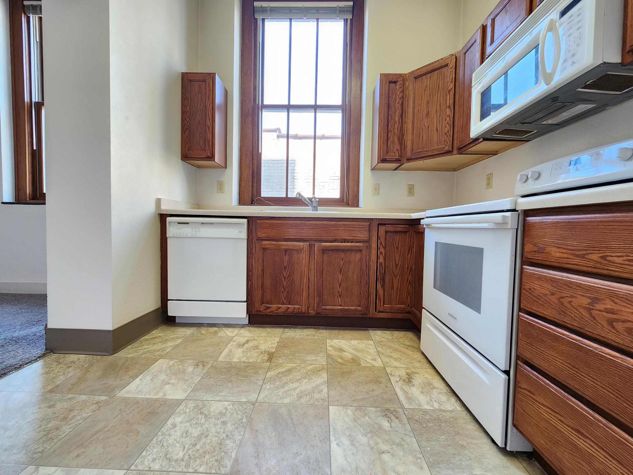 A kitchen featuring wooden cabinets, a dishwasher, and an oven with a stovetop. The countertop is light-colored, and there’s a large window allowing natural light to enter. The floor is tiled with a mix of cream and beige. The overall space is bright and inviting.
