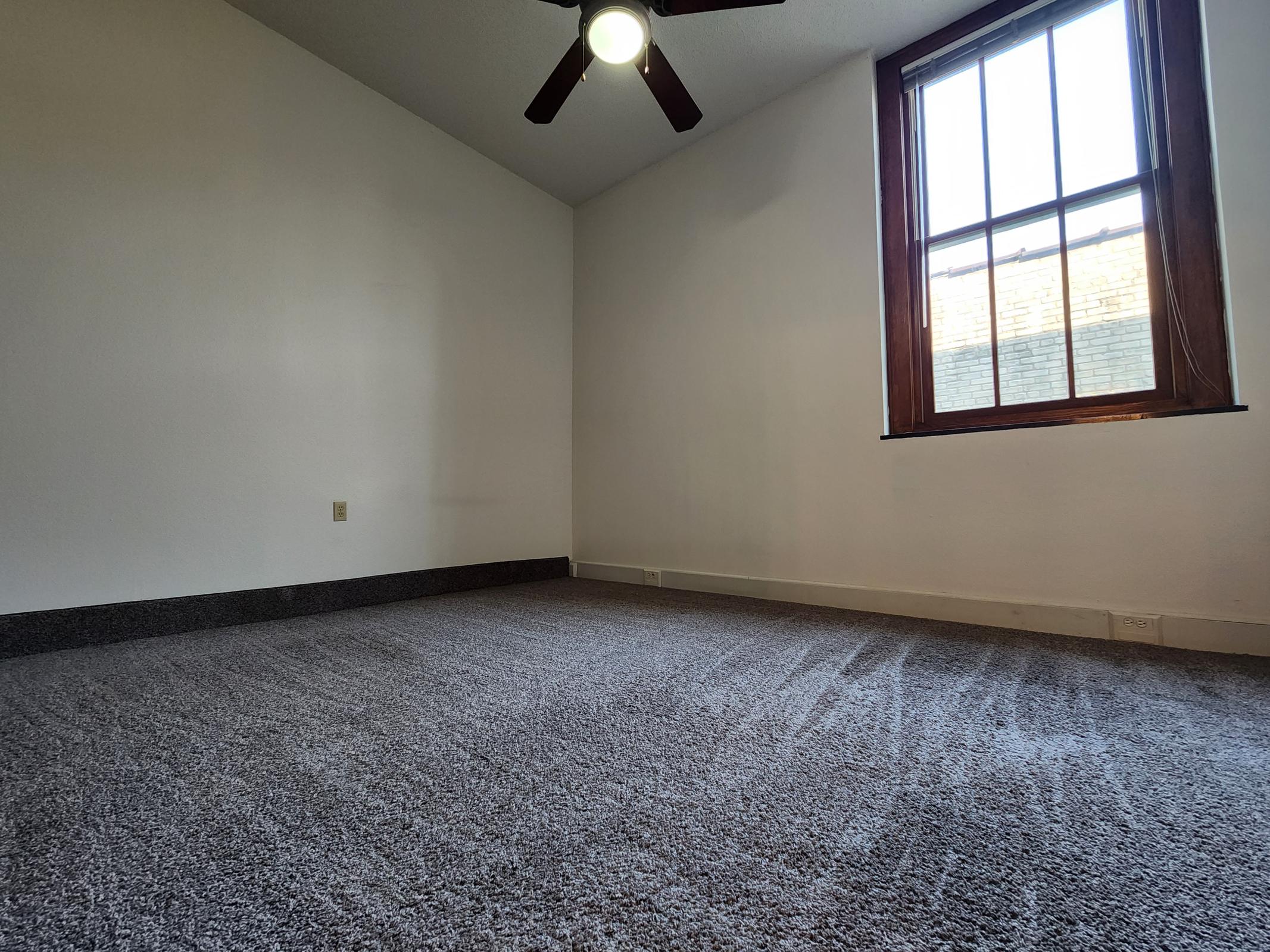 A spacious, empty room featuring beige carpet flooring and a white wall. A ceiling fan is mounted above, and a large window with wooden trim allows natural light to brighten the space. The room has a subtle, minimalist design, creating a sense of openness and potential for furniture arrangement.