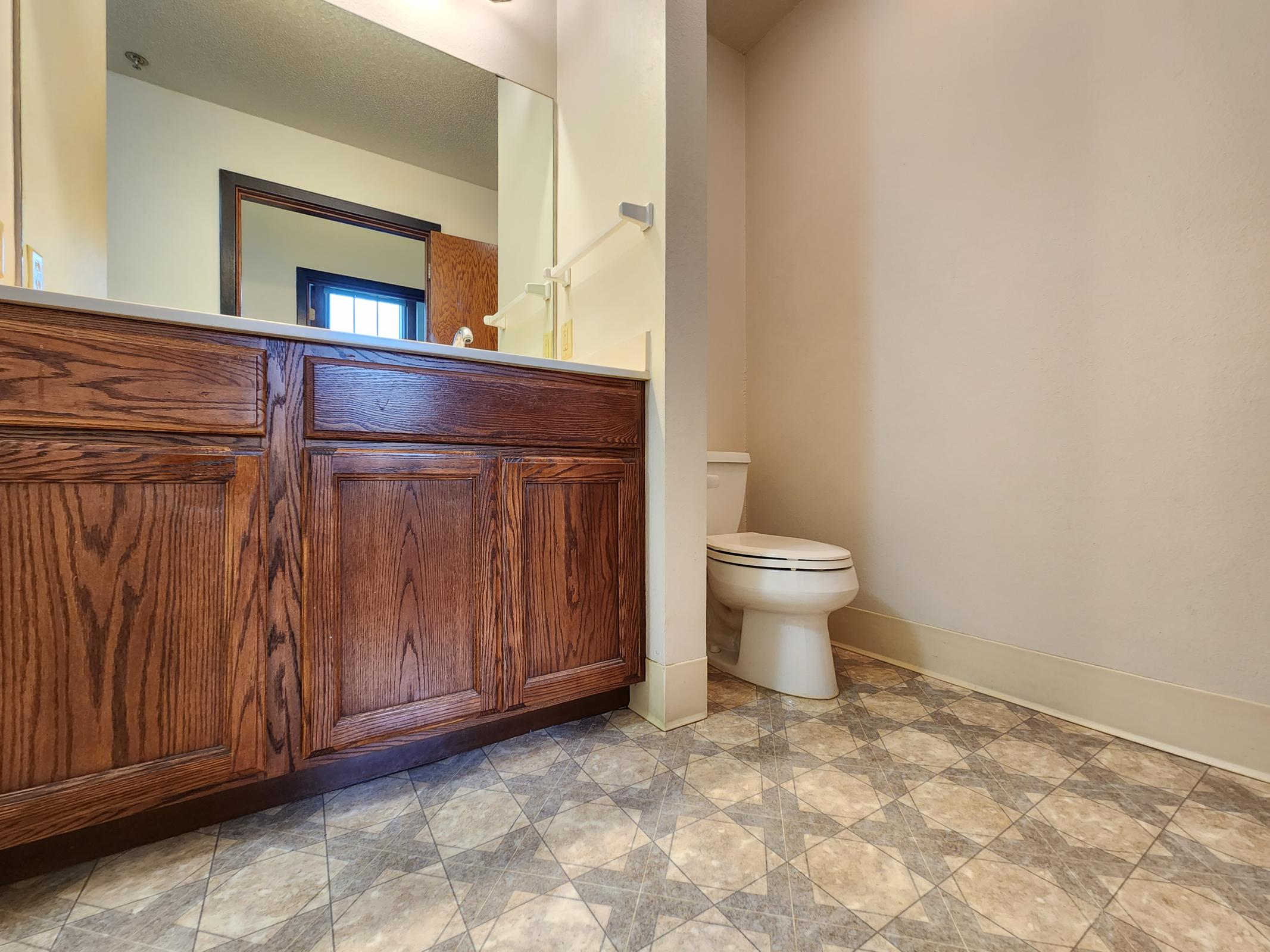 A bathroom featuring a wooden vanity with sink, a toilet, and tiled flooring. The walls are light-colored, and there's a mirror above the vanity. A door can be seen in the background, leading to an external area or hallway. The overall space is well-lit and functional, emphasizing a clean and simple design.