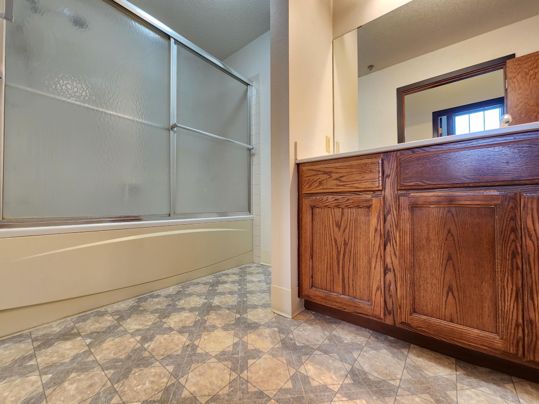 A bathroom featuring a bathtub and a glass shower enclosure. The vanity has wooden cabinets with a countertop. The floor is tiled in a diamond pattern with light and dark colors, and there is a door leading to another room in the background. Natural light is visible through a window.
