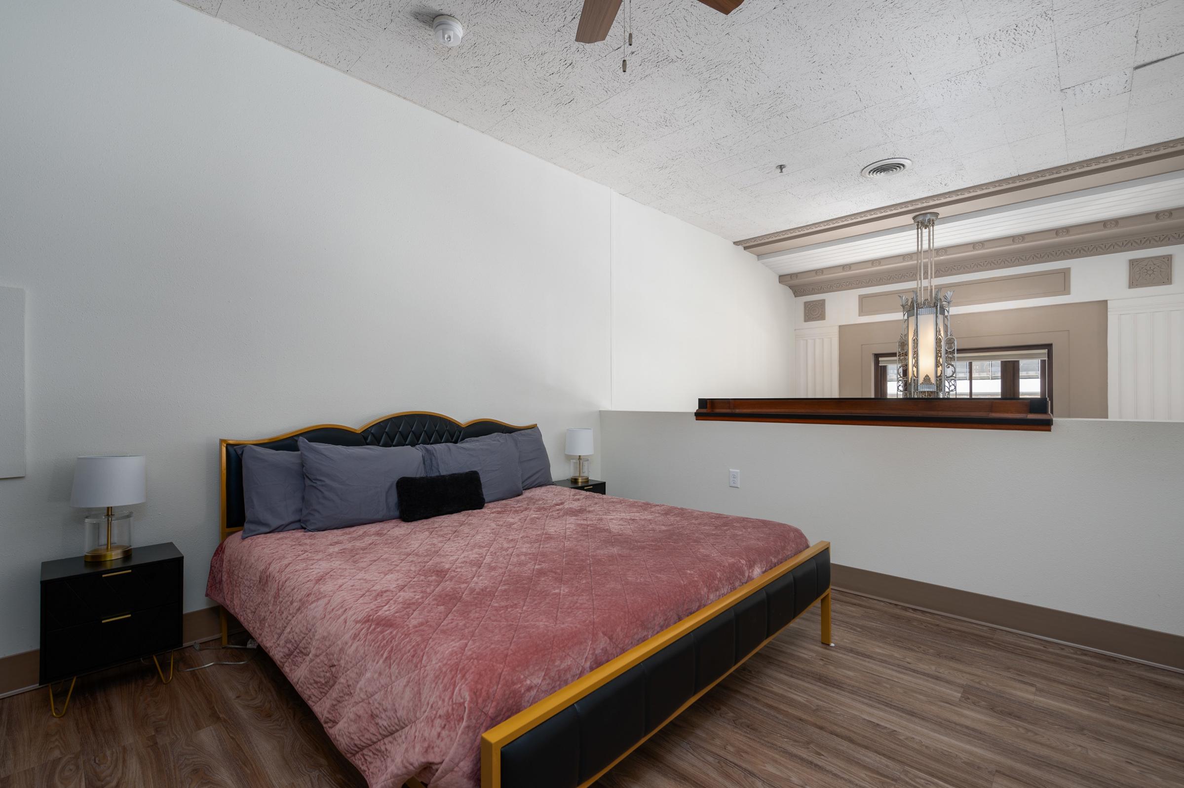 A modern bedroom featuring a black bed with a pink quilt, two bedside lamps on black nightstands, and a minimalist aesthetic. The room has light-colored walls, wooden flooring, and a decorative ceiling with a ceiling fan, creating a cozy and inviting atmosphere.