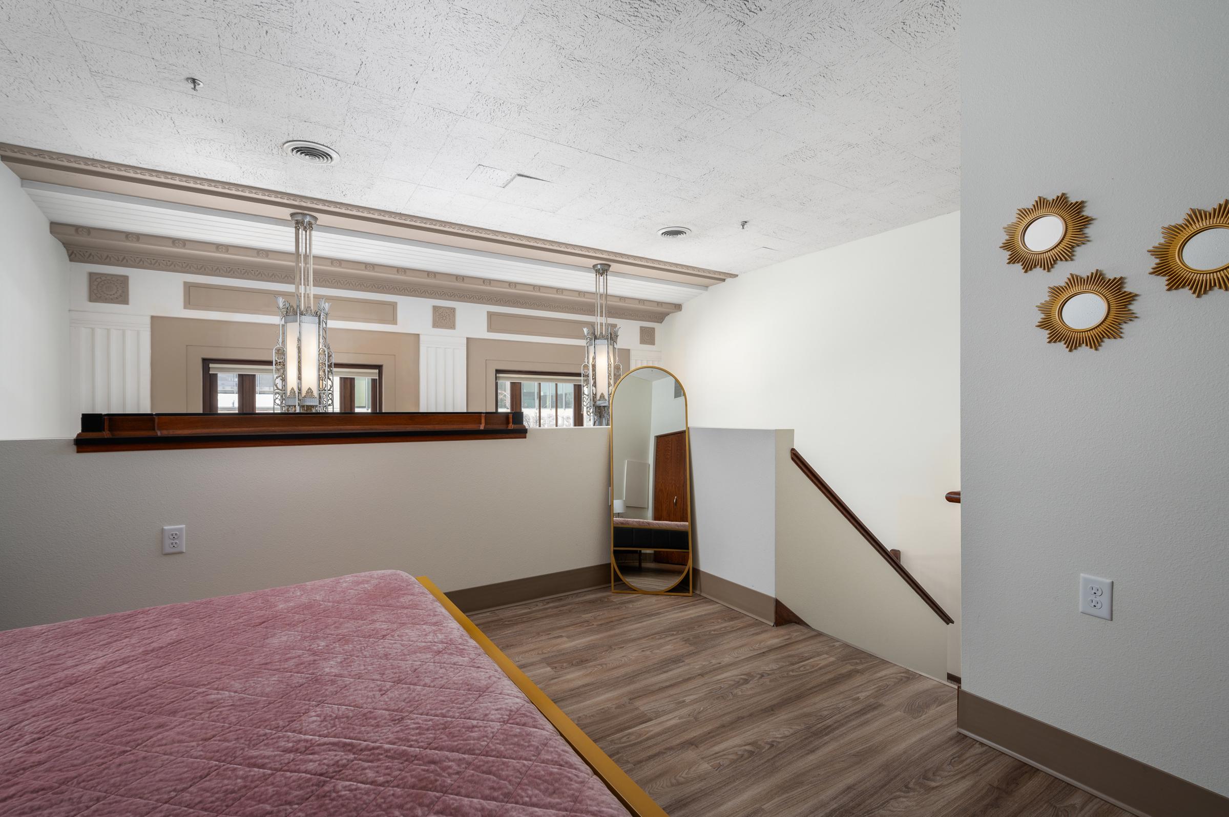 Interior view of a stylish room featuring a bed with a pink quilt, a round mirror, an elegant staircase, and decorative wall mirrors. The space showcases modern lighting fixtures and a blend of contemporary and classic architectural details, with wooden accents and a neutral color palette.