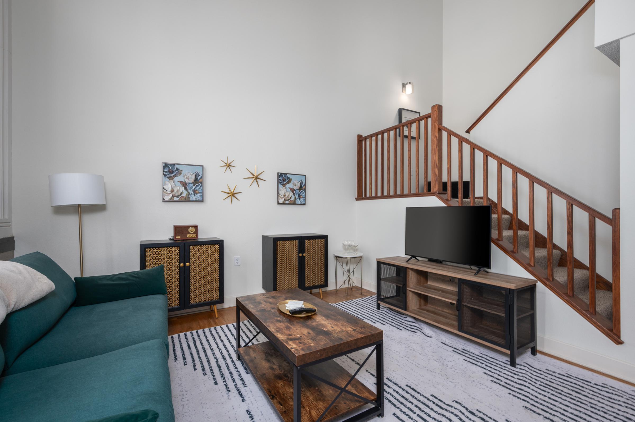 A cozy living room featuring a green couch, a wooden coffee table, and a TV stand. Two decorative cabinets are against the wall, adorned with framed artwork. A staircase with wooden railings leads to an upper level, and the space is illuminated by a wall fixture, creating a warm and inviting atmosphere.
