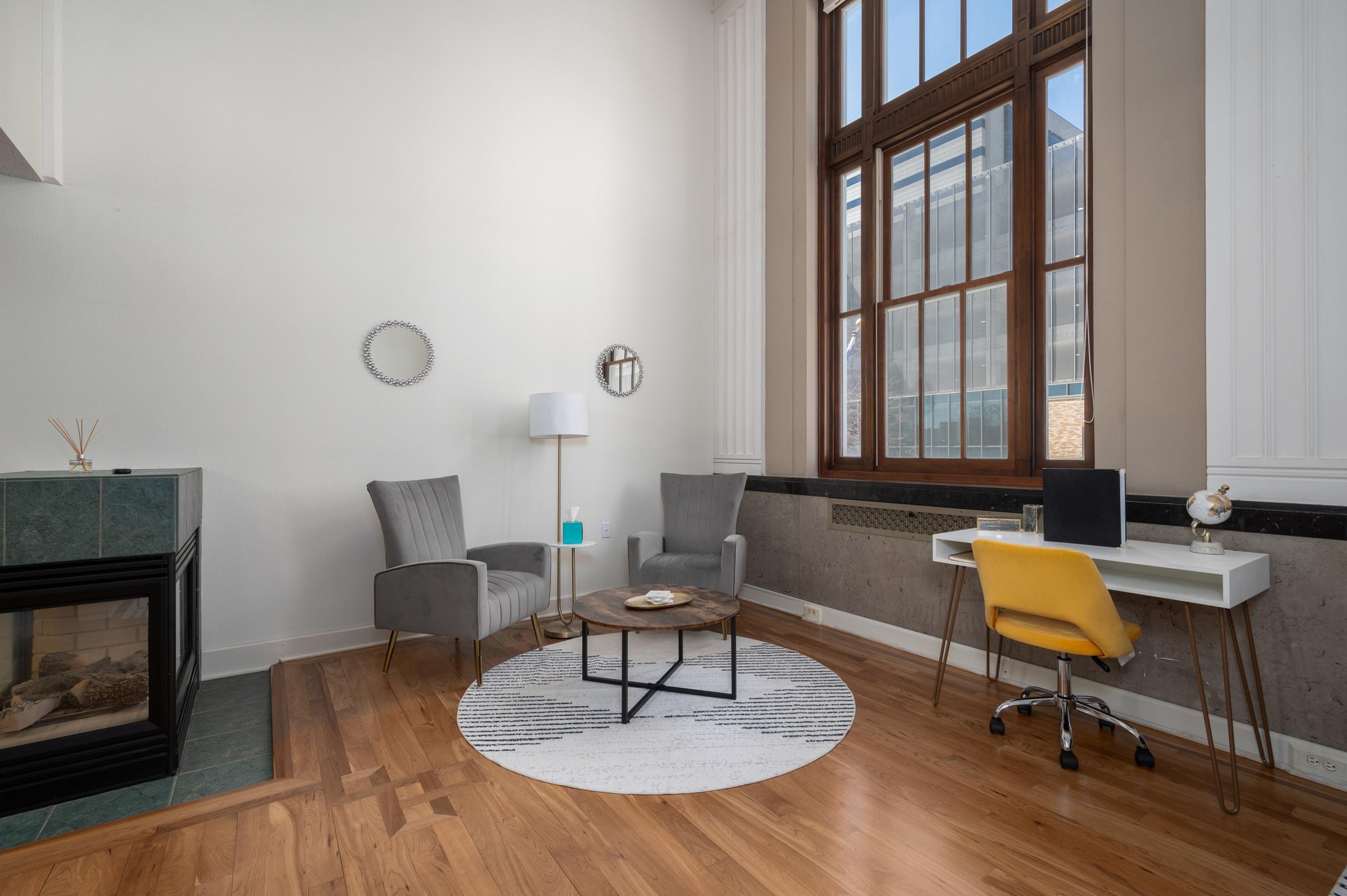 A cozy living area featuring two gray armchairs and a circular coffee table on a patterned rug. There is a modern white desk with a yellow chair and a lamp nearby. A large window lets in natural light, and a small mirror hangs on the wall, adding to the inviting atmosphere.