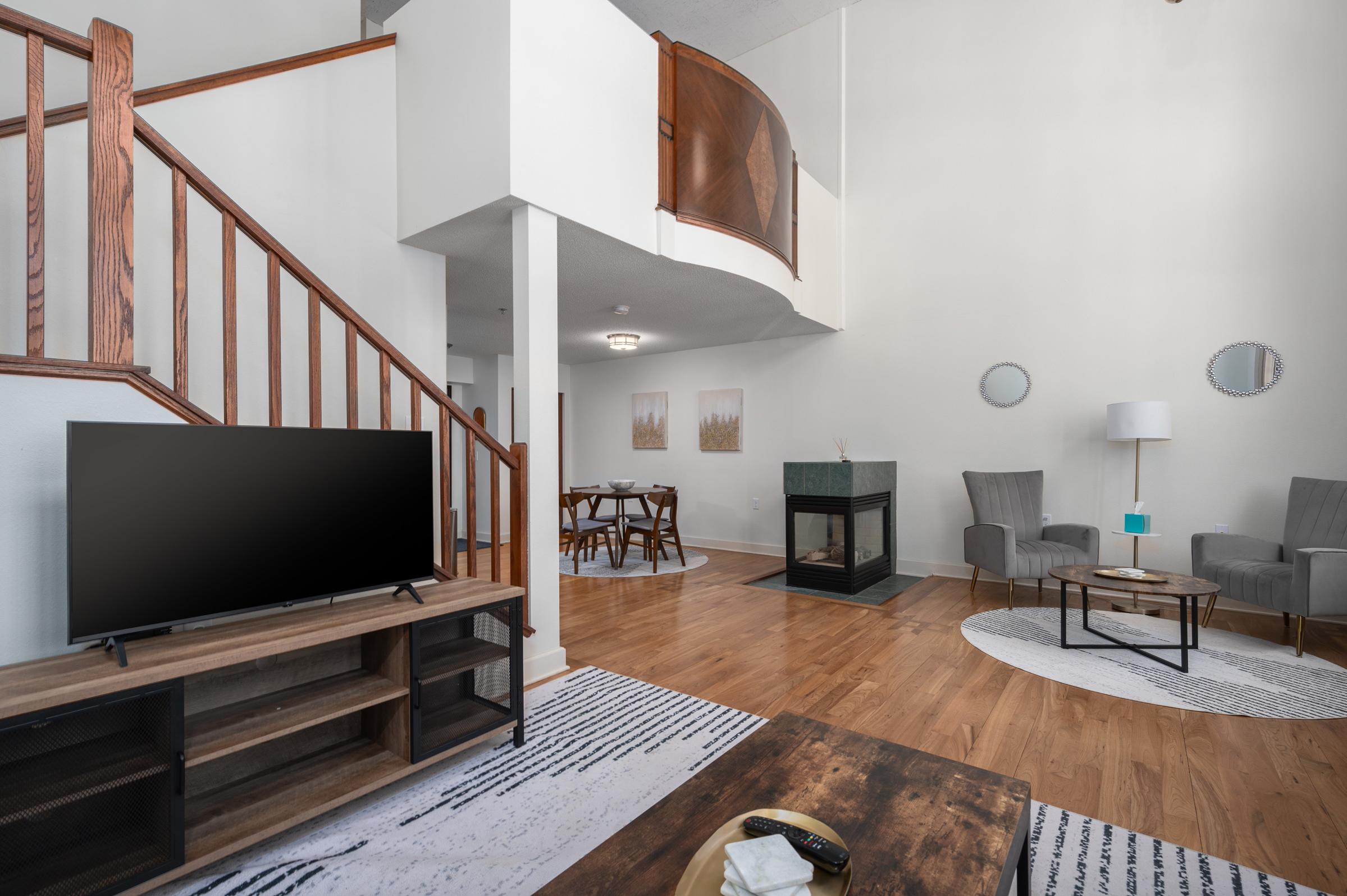 A modern living space featuring a wooden staircase, a large TV on a media console, a dining area with a round table, two gray chairs, and a decorative fireplace. The walls are painted in a light color, and there are circular rugs on the hardwood floor, creating a cozy atmosphere.