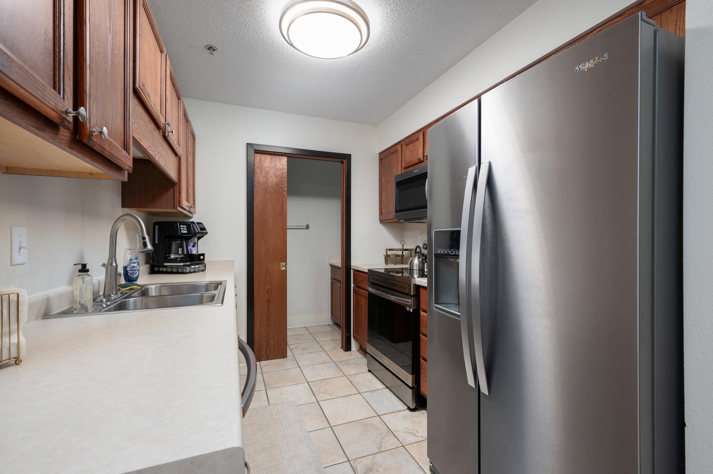 Modern kitchen featuring wooden cabinets, stainless steel appliances including a refrigerator and stove, and a sink. The countertop is light-colored, and tiled flooring adds to the contemporary design. A door leads to another room, and a coffee maker is visible on the counter.