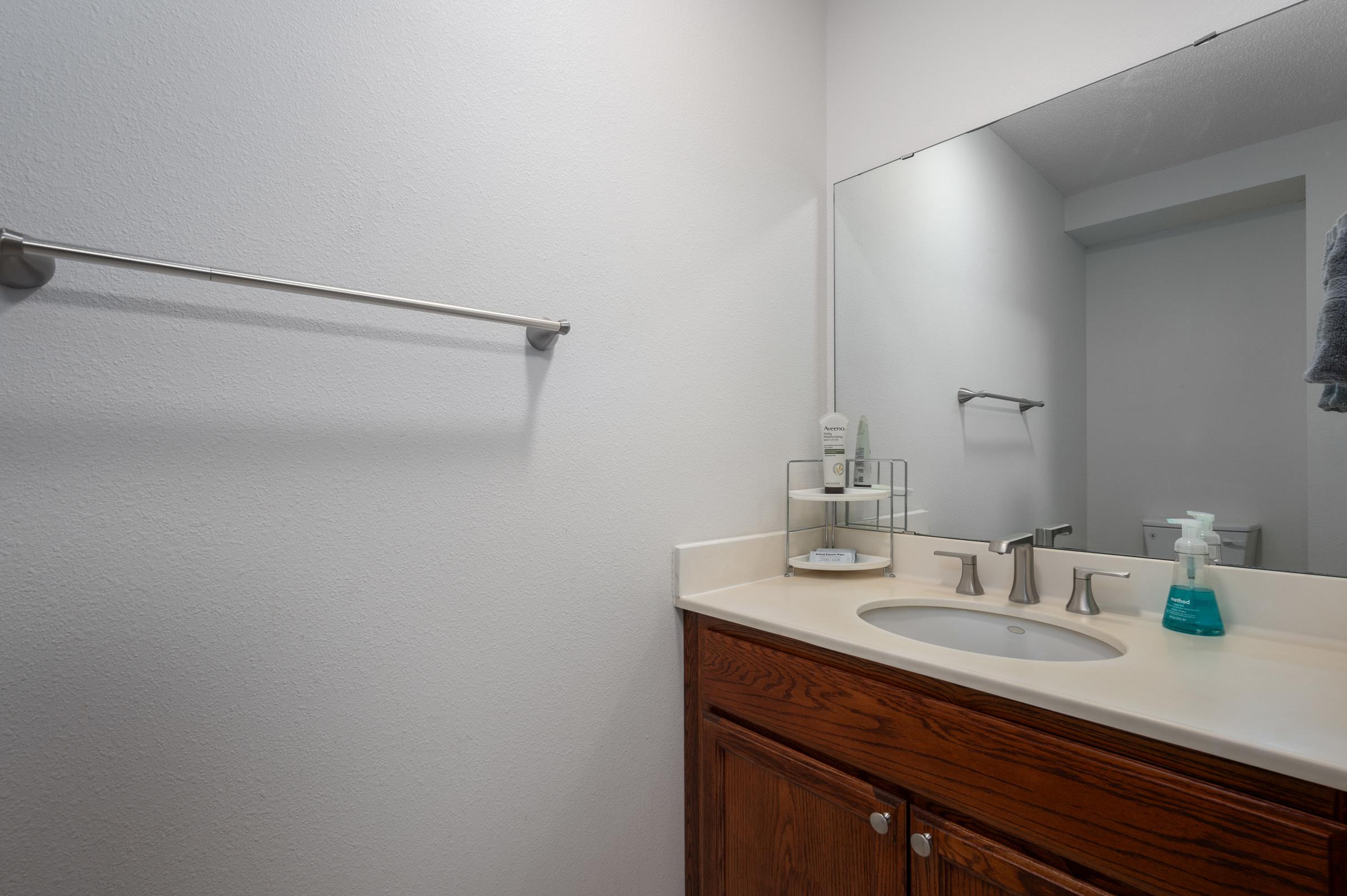 A clean and modern bathroom featuring a double sink with sleek faucets, a wall-mounted mirror, wooden cabinetry, and a shelf with toiletries. The walls are painted a light color, and there is a towel bar and a small container with hand sanitizer on the countertop.