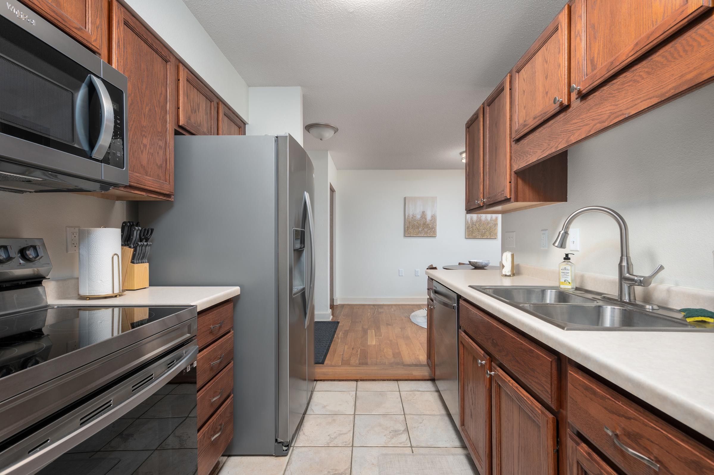 A modern kitchen with wooden cabinets and stainless steel appliances, including a microwave, refrigerator, and oven. The countertop is beige with a sink, and there are utensils neatly arranged. In the background, a doorway leads to a dining area with hardwood flooring and minimal decor.