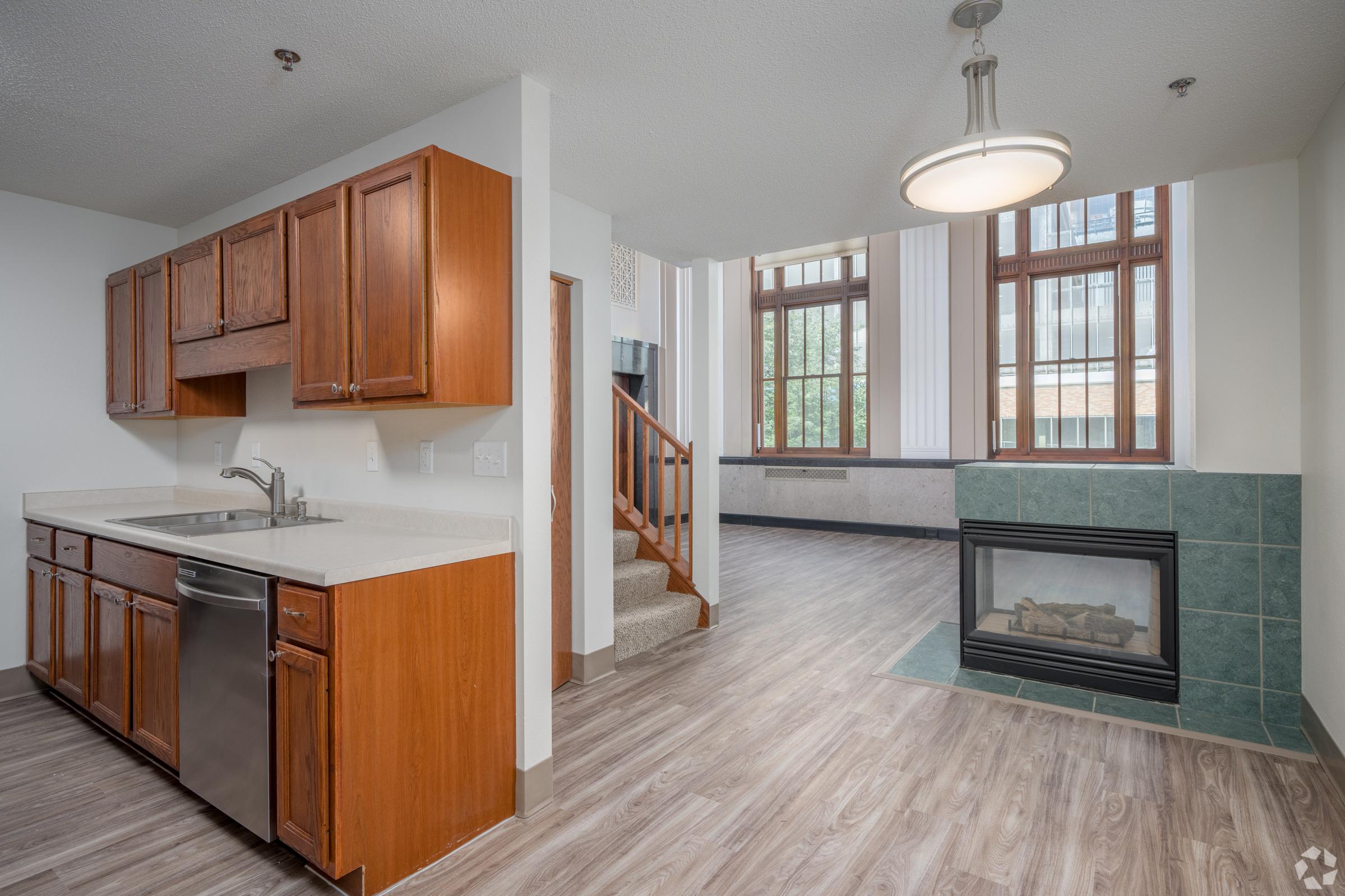 Bright, modern apartment interior featuring a kitchen with wooden cabinets, stainless steel appliances, and a countertop. The living area includes a gas fireplace and large windows, allowing natural light to fill the space. Stairs lead to an upper level, emphasizing an open layout.