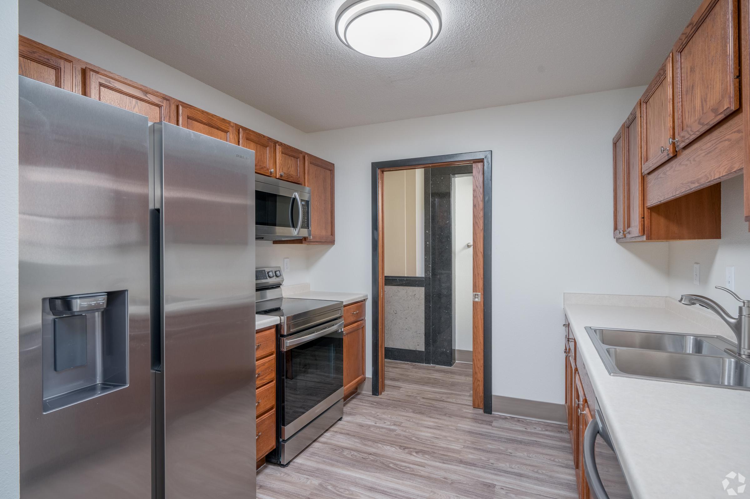 A modern kitchen featuring stainless steel appliances, including a refrigerator, microwave, and oven. The cabinetry is made of warm wood, complemented by a light-colored countertop. The floor is a light wood laminate, and there is a circular ceiling light enhancing the space. A doorway leads to another room in the background.