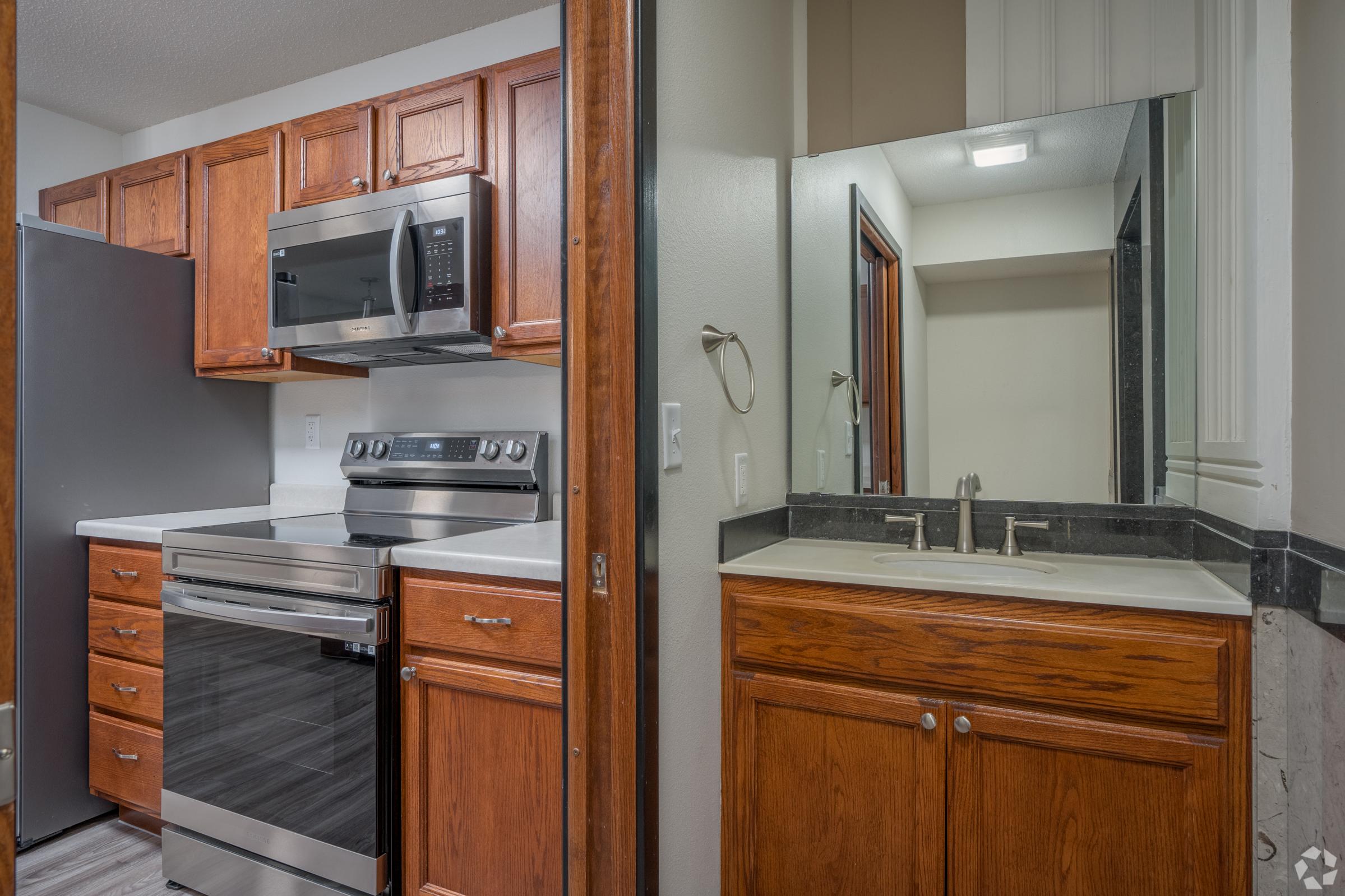 A modern kitchen featuring wooden cabinets, stainless steel appliances, and a countertop. There's a fridge, oven, and microwave on one side, and a bathroom vanity with a sink and mirror on the other, separated by a wall. The lighting is bright, creating a clean and inviting atmosphere.