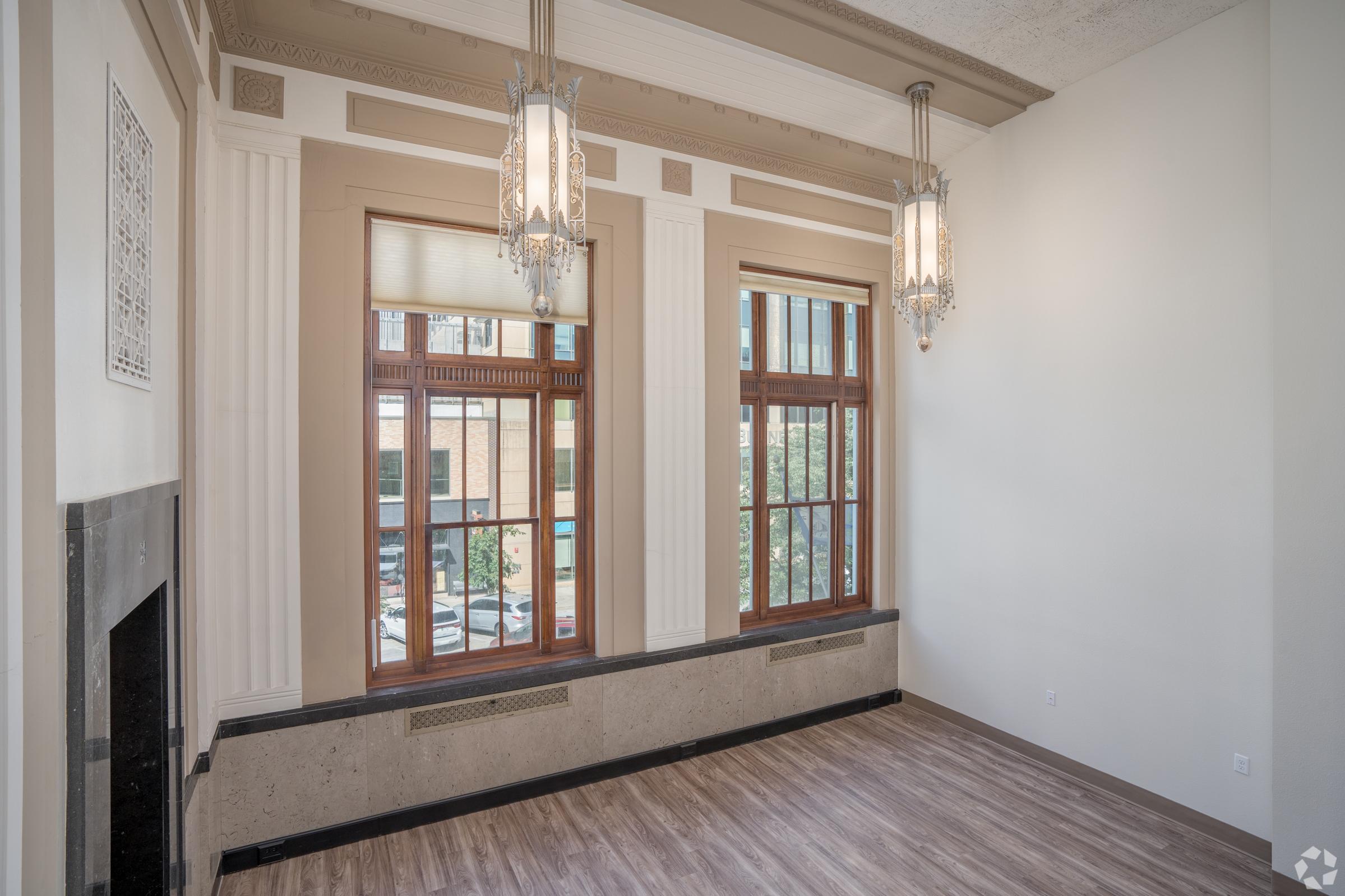 An empty room with large windows allowing natural light to enter. Elegant chandeliers hang from a high ceiling, and the walls feature decorative molding. A modern fireplace is visible, and the floor is covered with light-colored wood. The space has a clean, minimalist design.