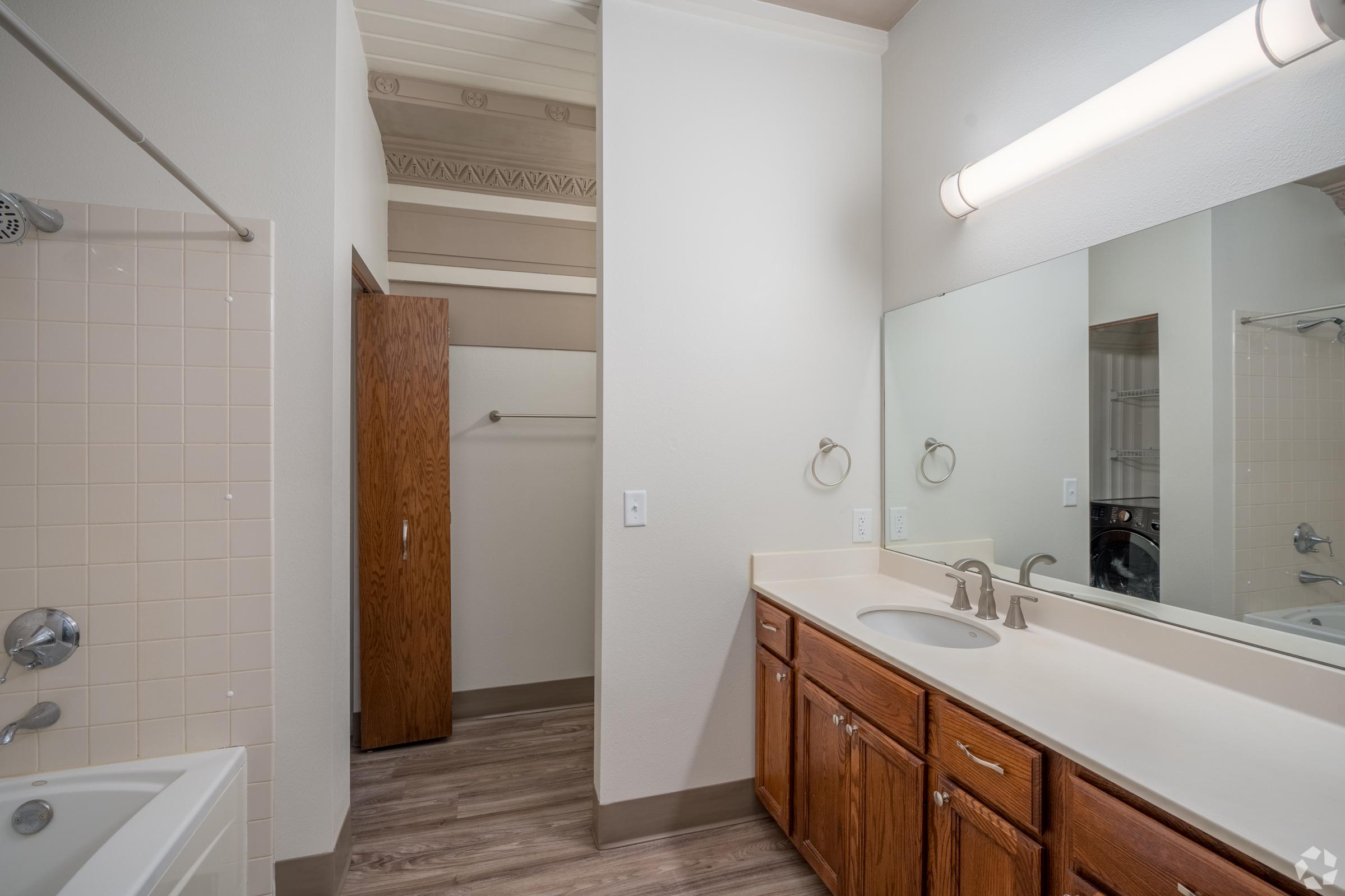 A clean and bright bathroom featuring a double sink vanity with wooden cabinets, a large mirror, and a bathtub. There is a shower curtain rod and a towel holder. The room has light-colored walls and wooden flooring, with a closet slightly visible in the background.