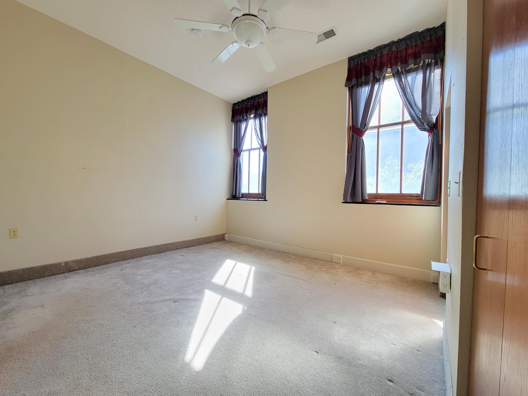 Empty room with light beige walls and carpeted floor. Two windows with sheer curtains allow natural light to enter, casting shadows on the floor. A ceiling fan is installed above. The overall atmosphere is bright and airy, with minimal decor.