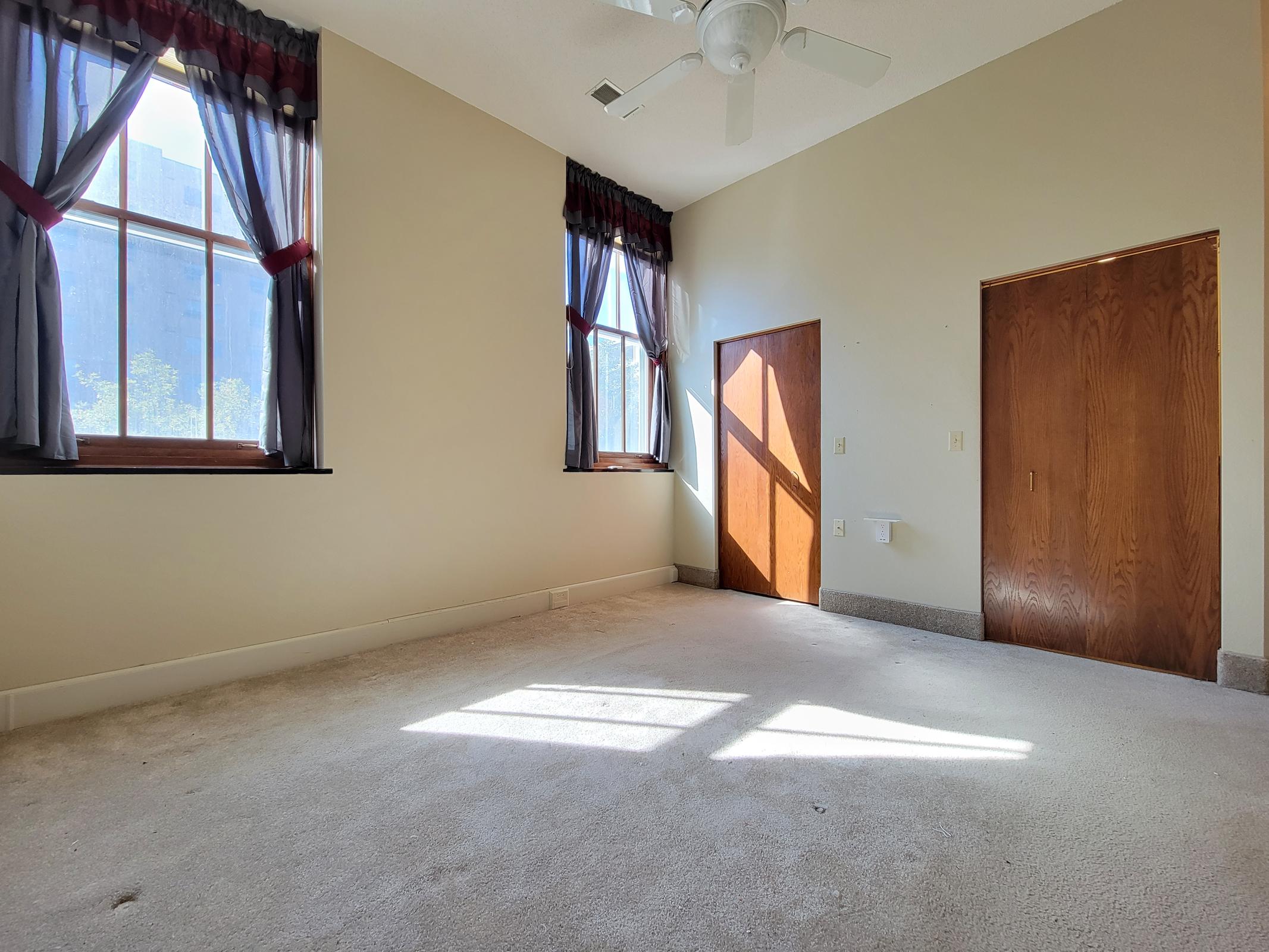 Empty room with light beige walls, a light-colored carpet, and large windows letting in natural light. There are two windows with red and black curtains, a ceiling fan, and two wooden doors on the right side of the room. The floor is bare except for some shadows created by the light coming through the windows.