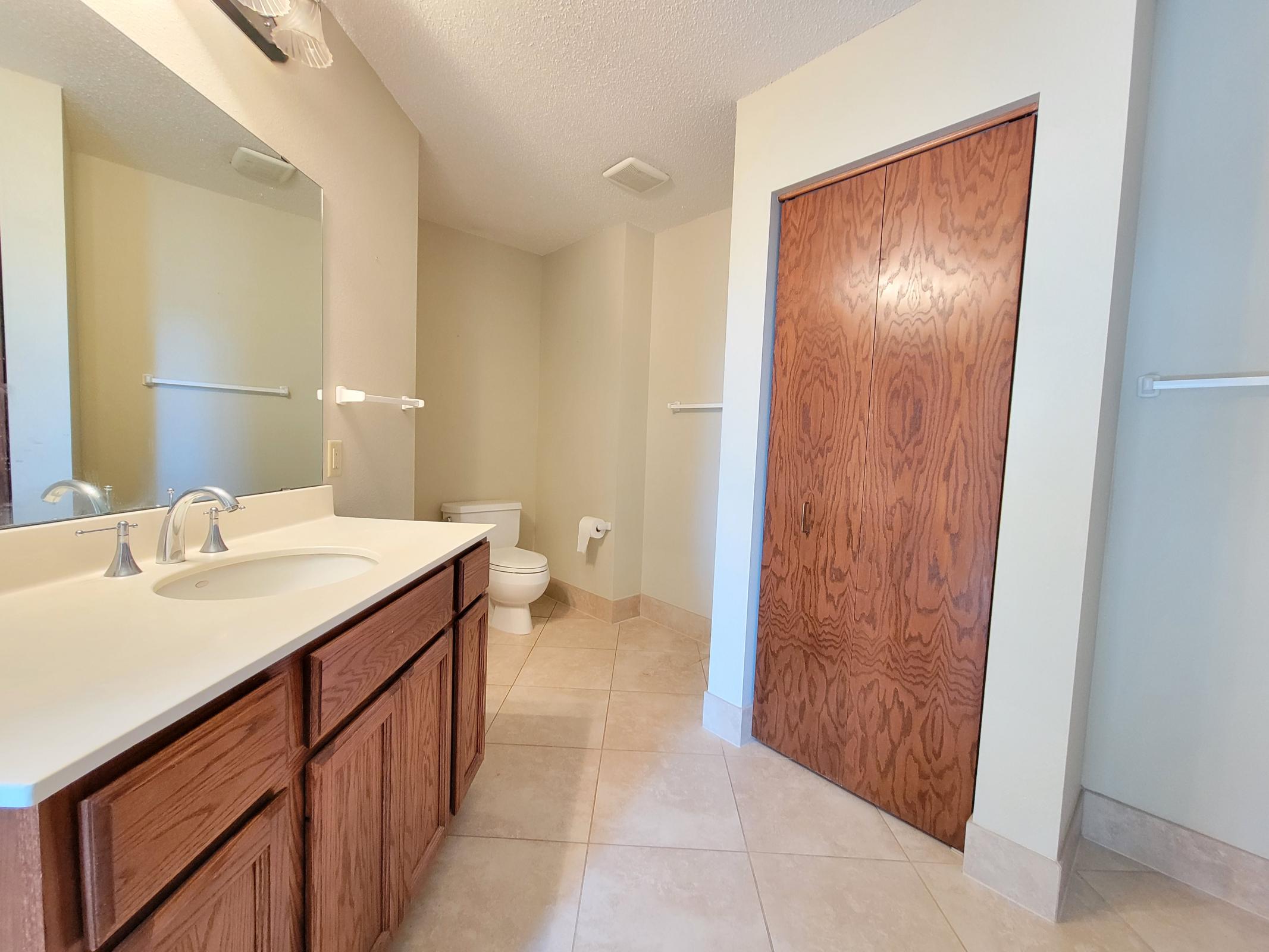 A clean and spacious bathroom featuring a double sink vanity with wooden cabinetry, a large mirror, and a toilet. The floor is tiled, and there's a wooden door leading to a storage area. The walls are painted in a light color, enhancing the openness of the space.