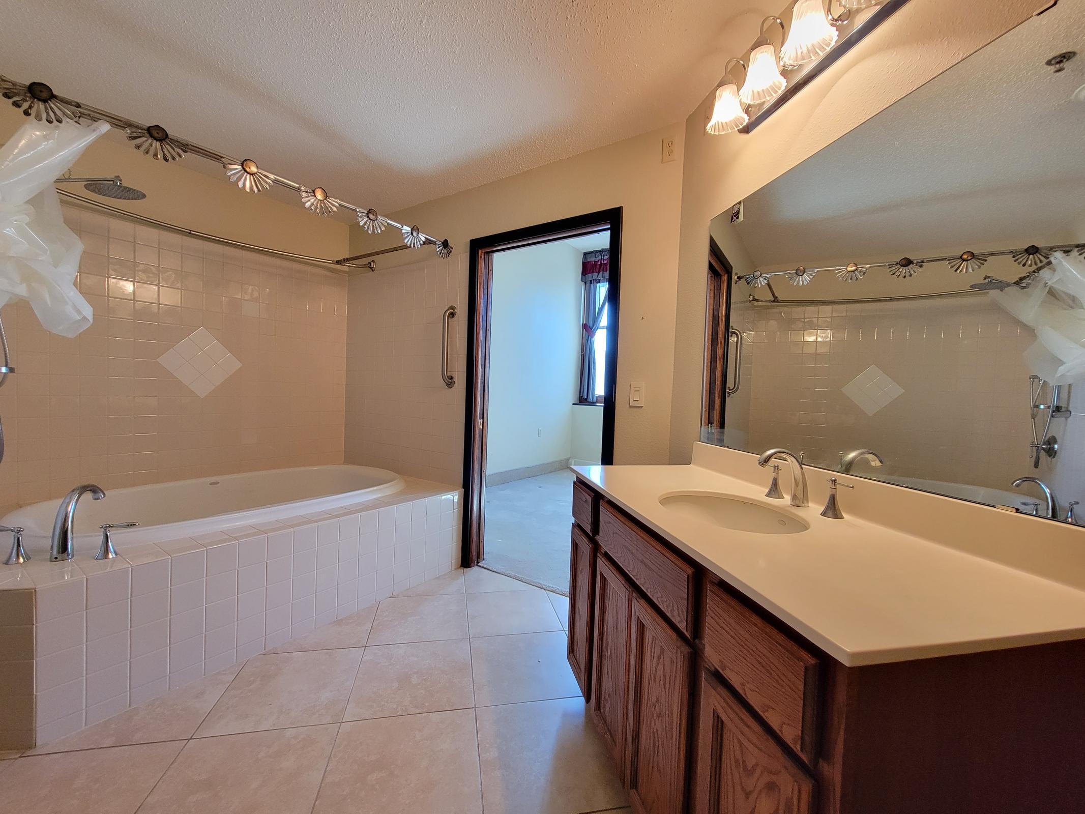Spacious bathroom featuring a large soaking tub, a separate shower area, a double sink vanity with wooden cabinets, and a large mirror. The room has neutral tile flooring and walls, with natural light coming through a window. A towel rack and a decorative shower curtain are also visible.