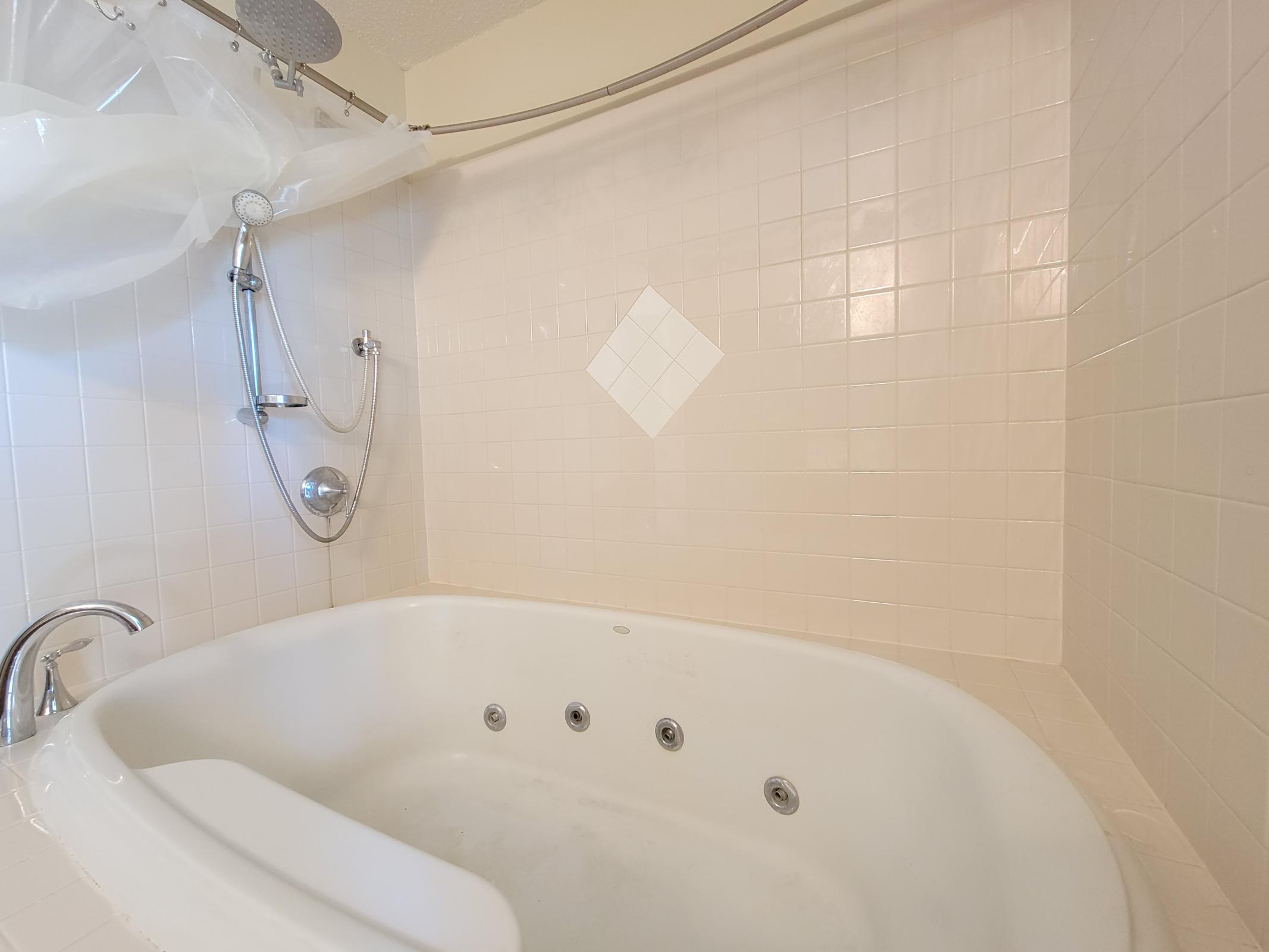 A clean bathroom featuring a well-maintained bathtub with jet fixtures. The surrounding walls are covered in light beige tiles, and a showerhead is installed above with a plastic curtain hanging. The space is bright and uncluttered, creating a serene atmosphere.