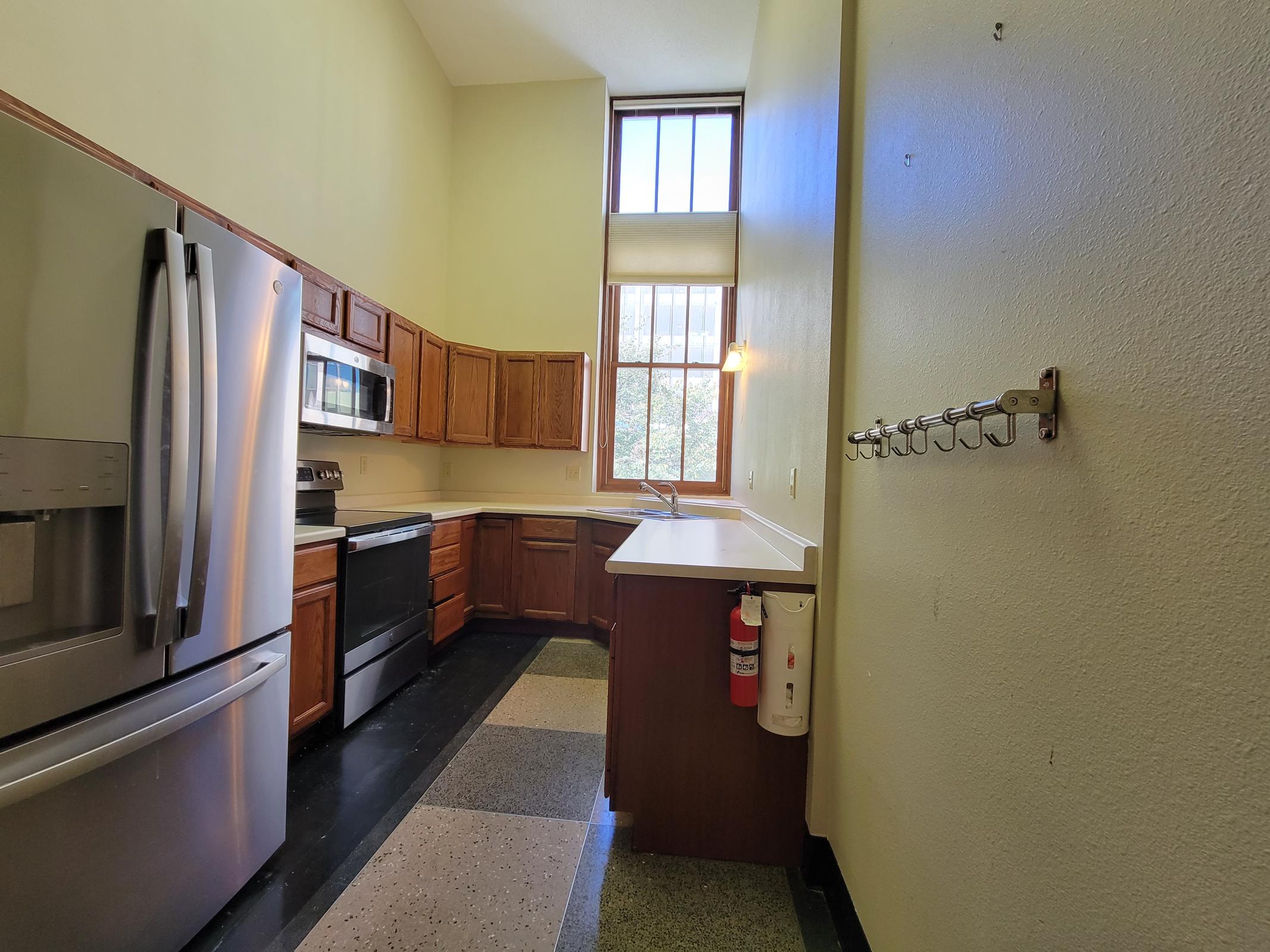 A spacious kitchen featuring wooden cabinets, stainless steel appliances, a large window allowing natural light, and a black and grey tiled floor. There is a wall with hooks and a fire extinguisher mounted nearby.