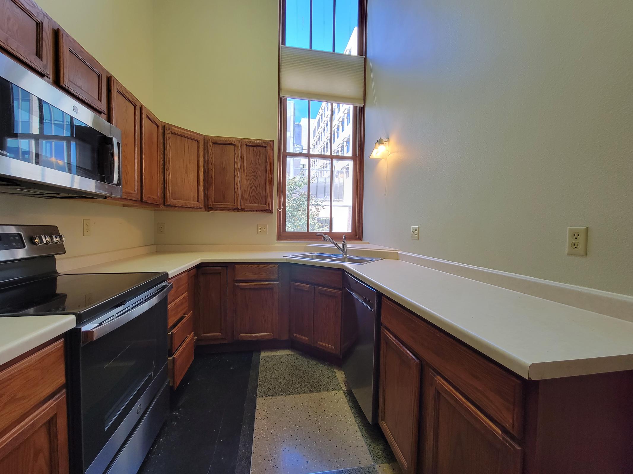 A bright kitchen featuring wooden cabinets, a modern stainless steel stove and dishwasher, and a double sink. The countertops are light-colored, and there is a large window letting in natural light, offering a view of the exterior. The flooring has a mix of tiles and concrete.