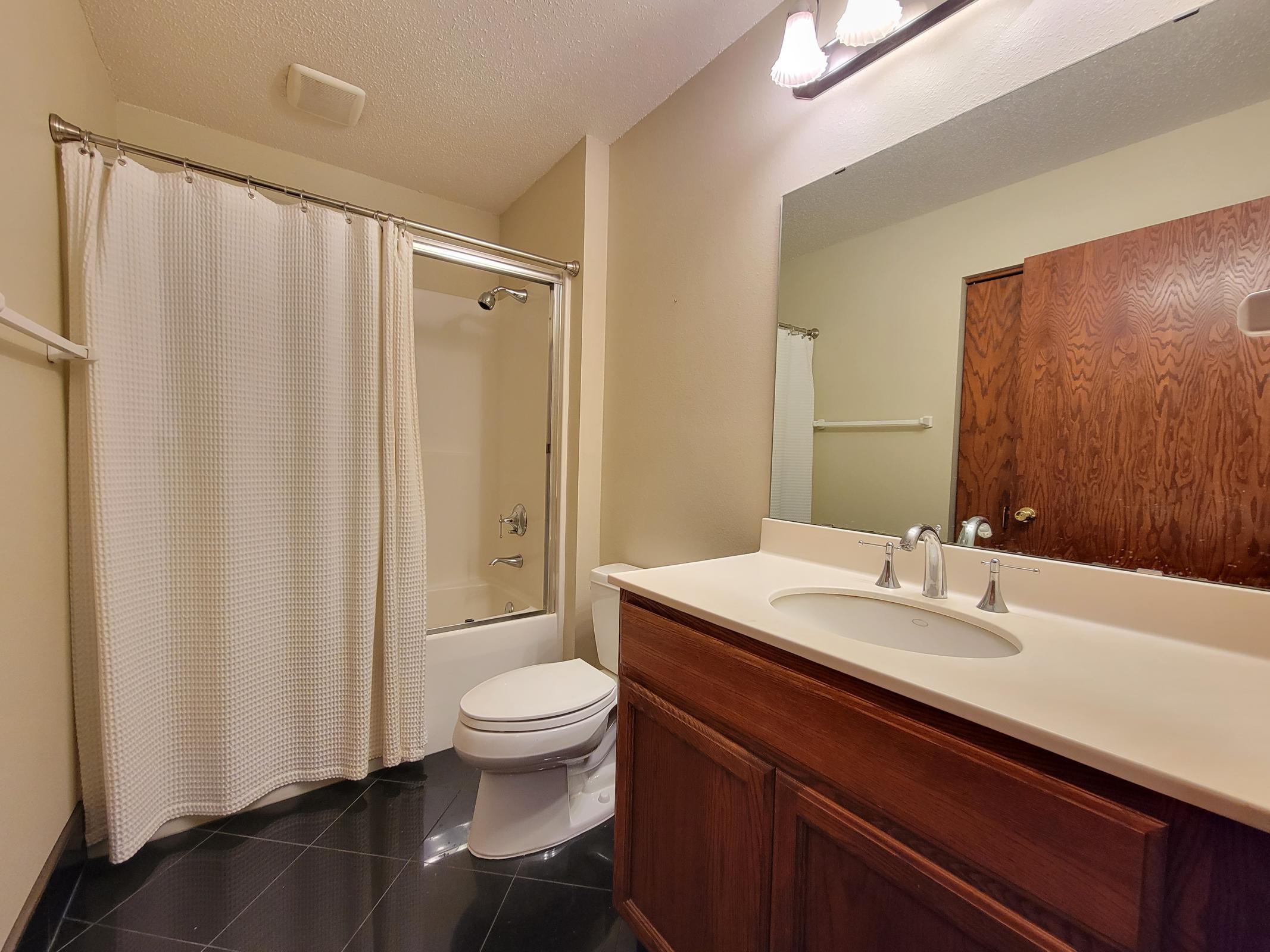 A bathroom featuring a shower with a white curtain, a modern sink with a countertop, a toilet, and dark tiled flooring. A mirror above the sink reflects the space, and a wooden door is visible in the background. Natural light seems to illuminate the area, creating a clean and spacious atmosphere.
