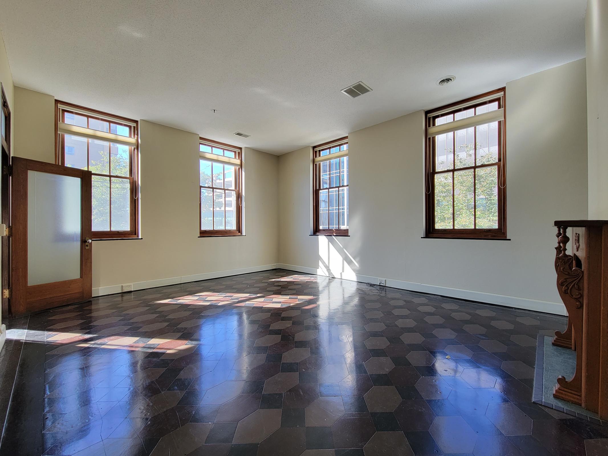 Spacious room with large windows allowing natural light to fill the space. The floor features a unique hexagonal pattern, and the walls are painted in a light color. There is a decorative wooden feature on one side, enhancing the room's character. The atmosphere is bright and inviting.