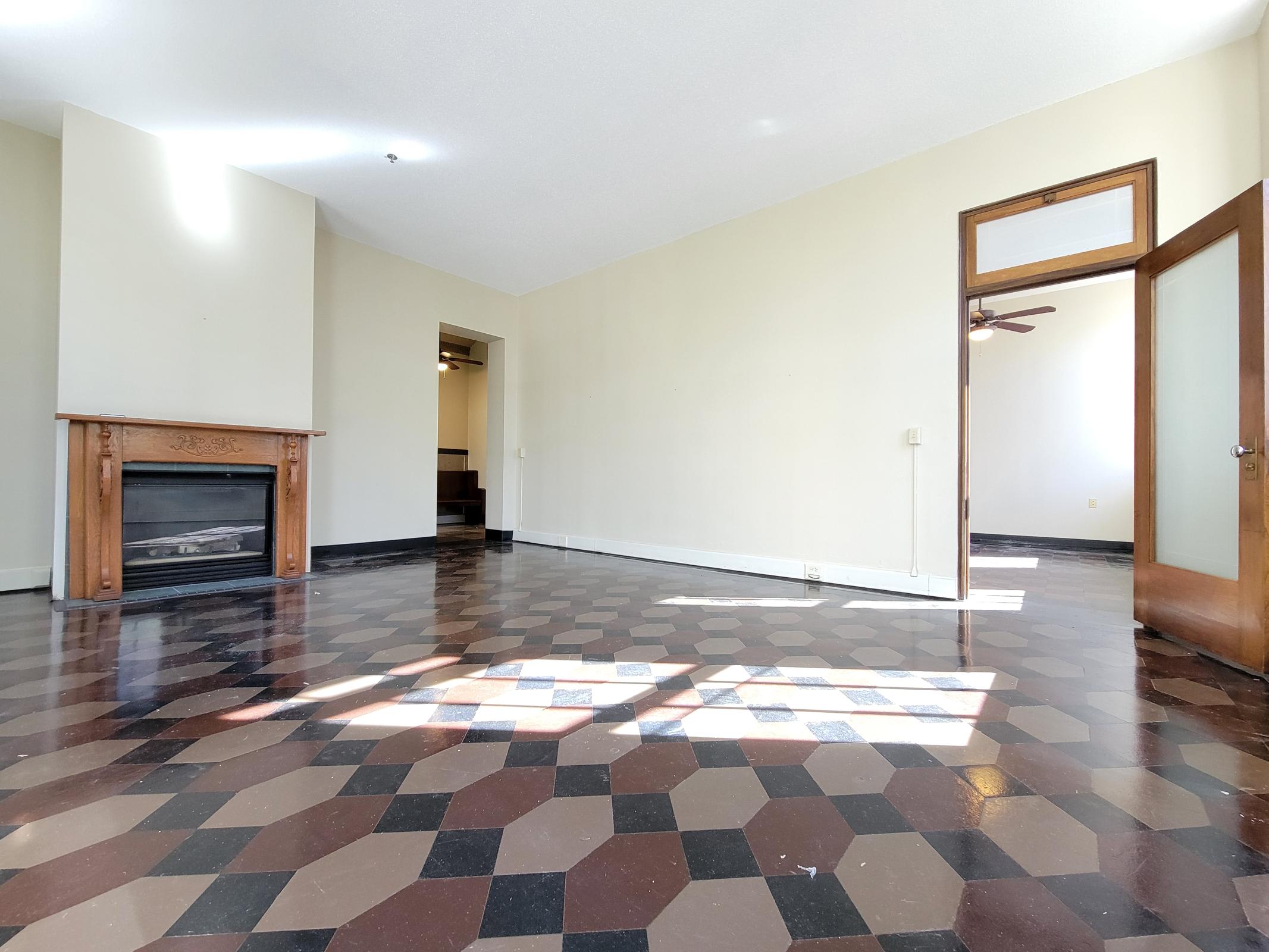 A spacious, empty living room featuring a decorative fireplace, hardwood flooring with a geometric pattern, and large windows allowing natural light. The walls are painted a soft neutral color, and there is a door leading to another room in the background. The overall ambiance is bright and open.
