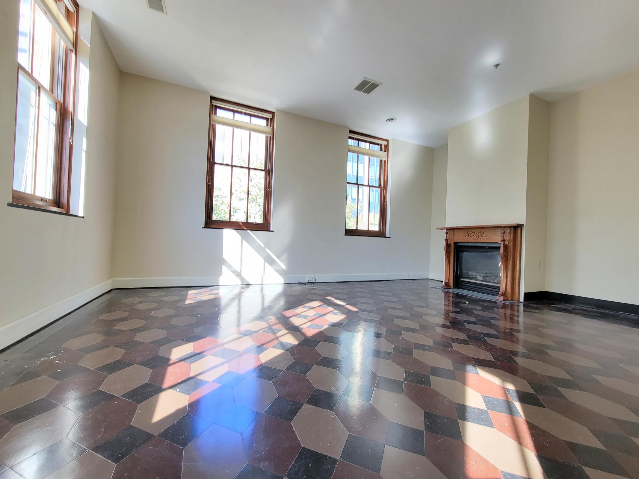 Spacious room with large windows allowing natural light to fill the space. The flooring features a unique hexagonal pattern in shades of brown and black, with a central area near a fireplace. The walls are painted in a neutral color, creating a bright and open atmosphere.