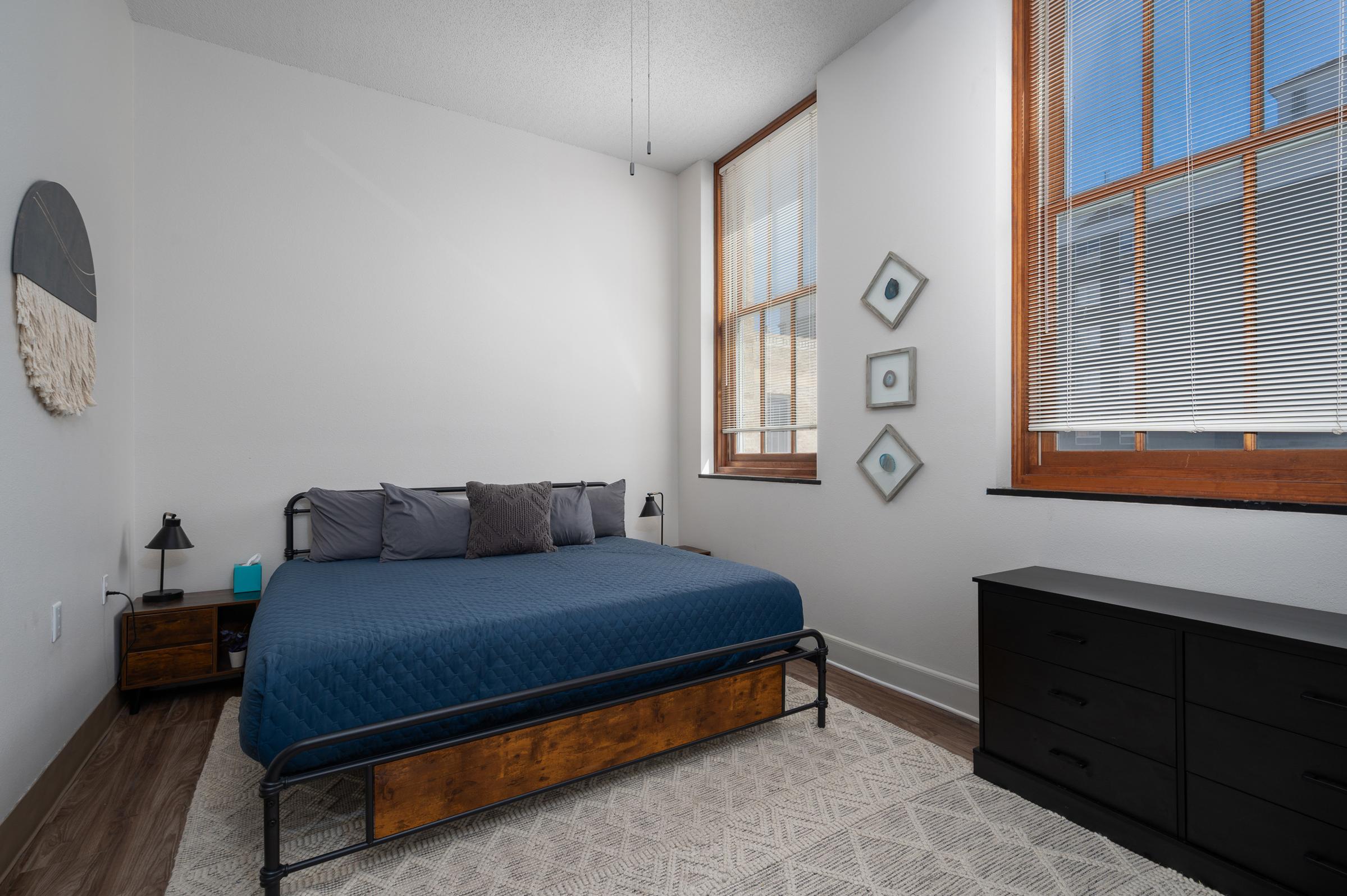 A modern bedroom featuring a blue quilted bed with several pillows, a wooden nightstand with a lamp, and a sleek black dresser. Large windows allow natural light to illuminate the room, which has light-colored walls and decorative wall art. The flooring is a warm-toned laminate, enhancing the cozy atmosphere.