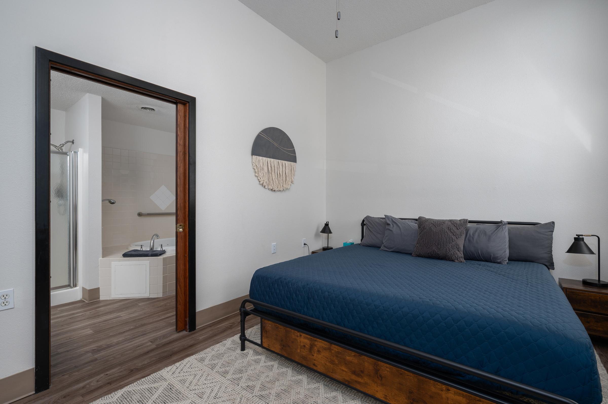 Modern bedroom featuring a dark blue bedspread on a stylish metal bed frame. There are two bedside lamps and a textured area rug. A doorway leads to a bathroom equipped with a shower and bathtub, visible in the background. The walls are painted white, enhancing the spacious feel of the room.