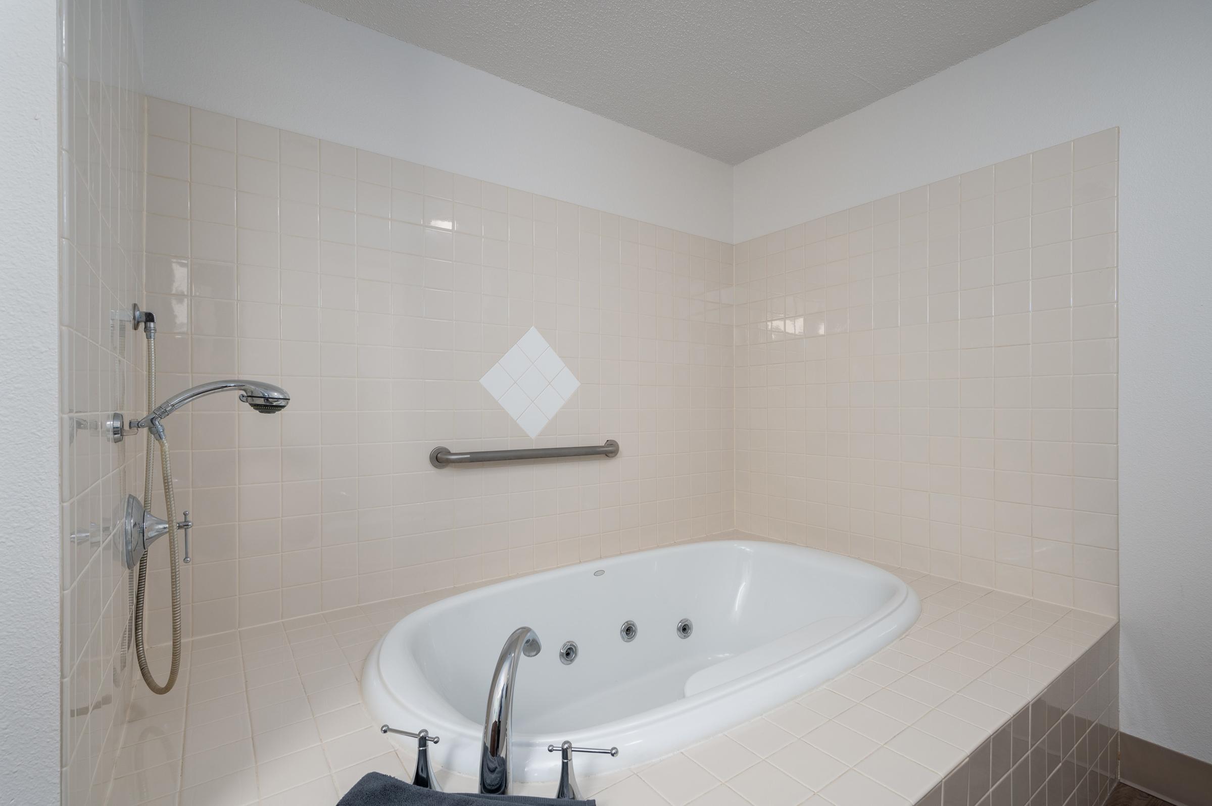 A spacious, mostly white bathroom featuring a large, oval jacuzzi tub with a shower head attached to the wall, a grab bar for safety, and tiled walls. The floor is light brown tile, and the overall space has a clean, modern aesthetic.